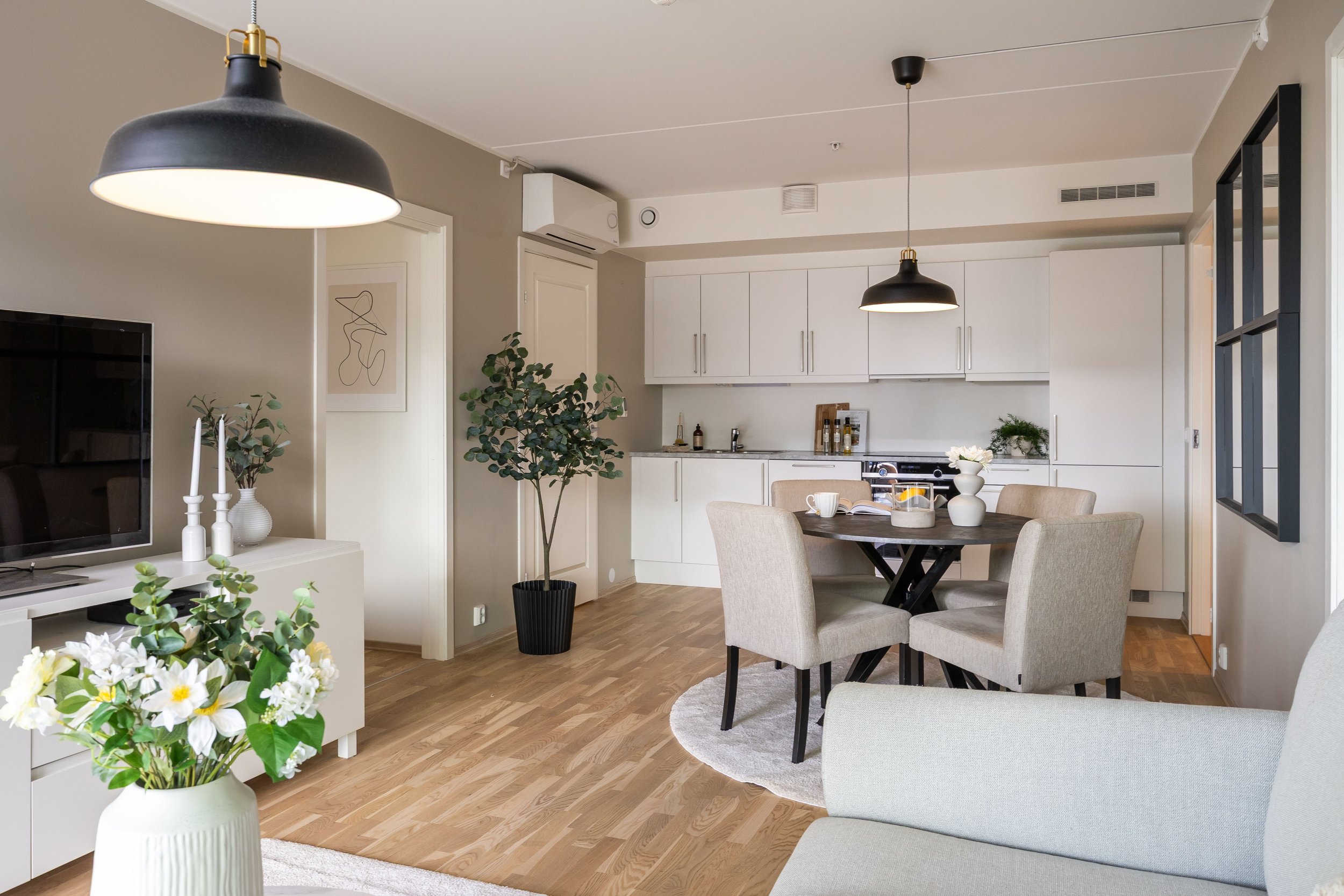 Et moderne, lyst kjøkken og spiserom med hvite kjøkkenskap, et rundt bord med fire stoler, grønne planter, og to hengende taklamper over bordet. Det er også en TV og dekorative blomster på en hylle.