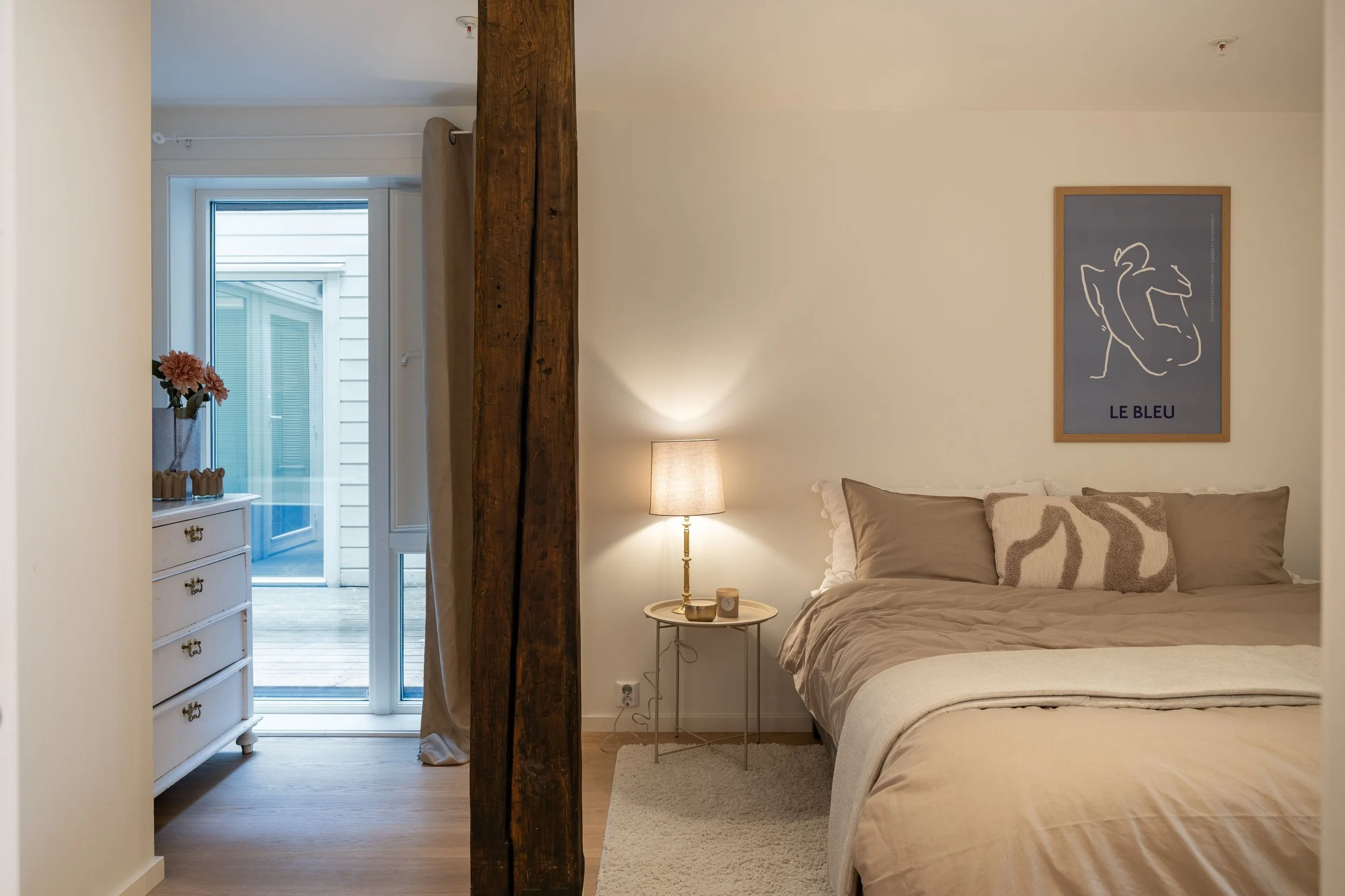 Et soverom med seng, et bilde på veggen med teksten 'LE BLEU', et nattbord med en lampe og dekorasjon, en garderobe, og en utgang til en balkong eller terrasse. Det er en vertikal bjelke som deler rommet.