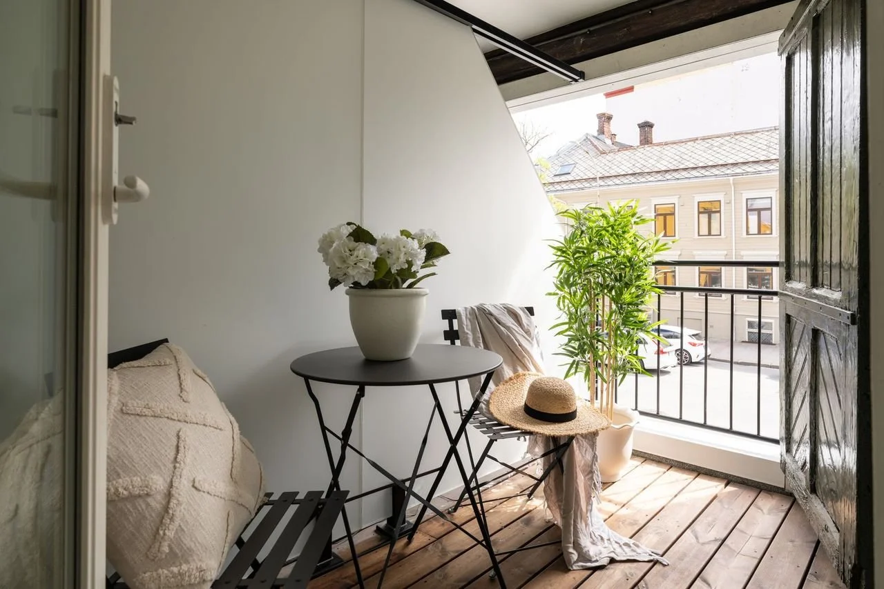 En liten balkong med tre møbler, en plantestand med stor plante, og en stol med et stråhatt på. Utsikt over en gate og en bygning med flere vinduer.