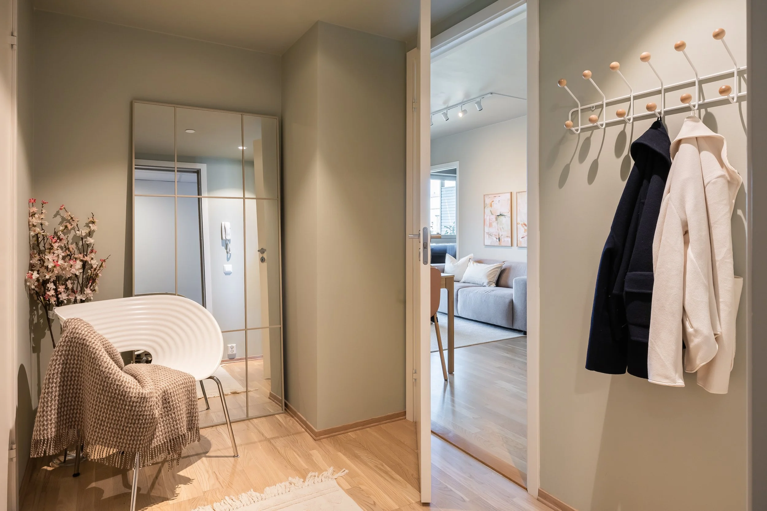 Innendørs utsikt gjennom en dør mot en stue med sofa og malerier, og en entré med speil, stol, og krokhenger med klær.
