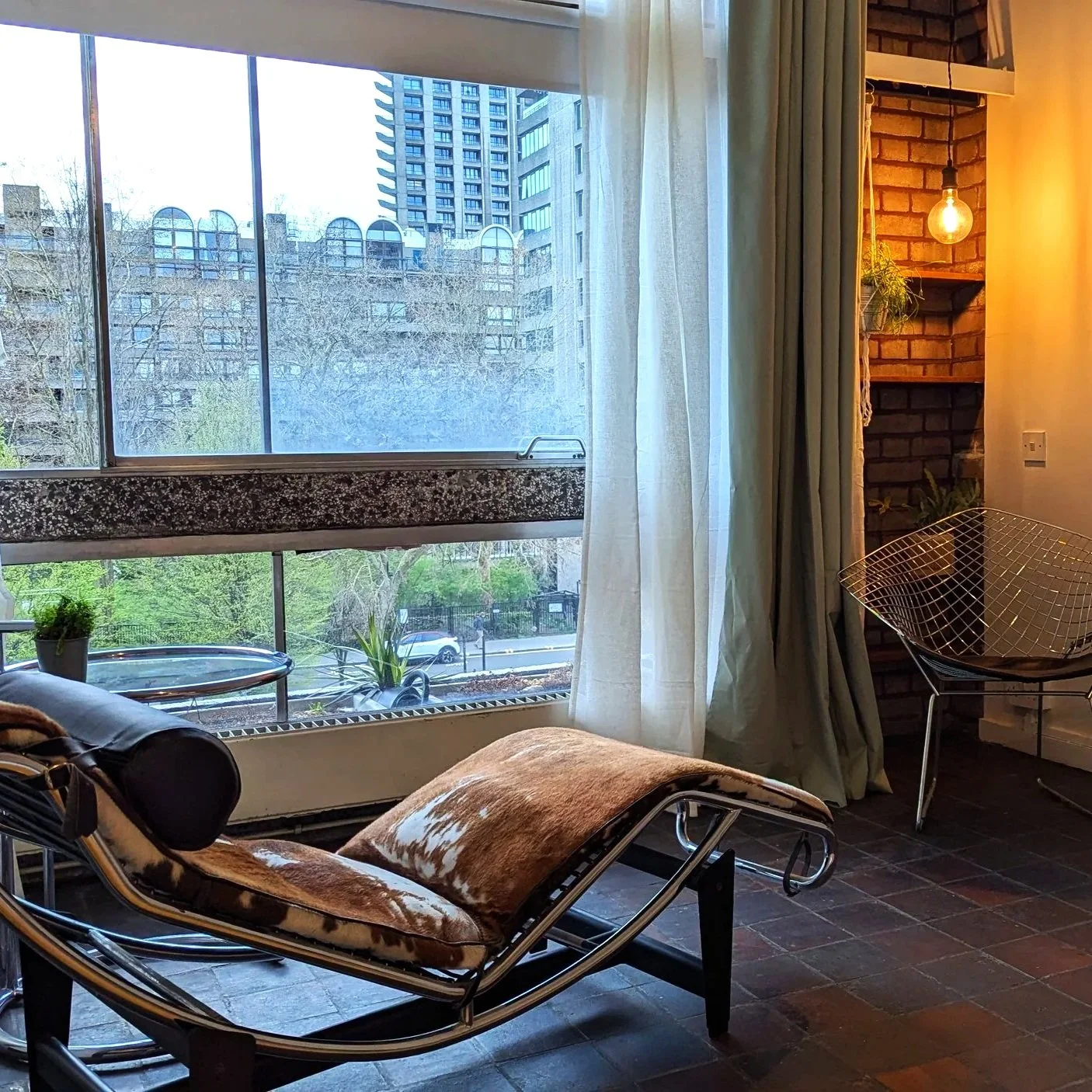 Interior of a post war brutalist era building , modern living room with a large window showing city buildings. A famous cassina 4 chaise lounge, and a small side table with a  wire chair, and warm lighting with exposed brick wall features.