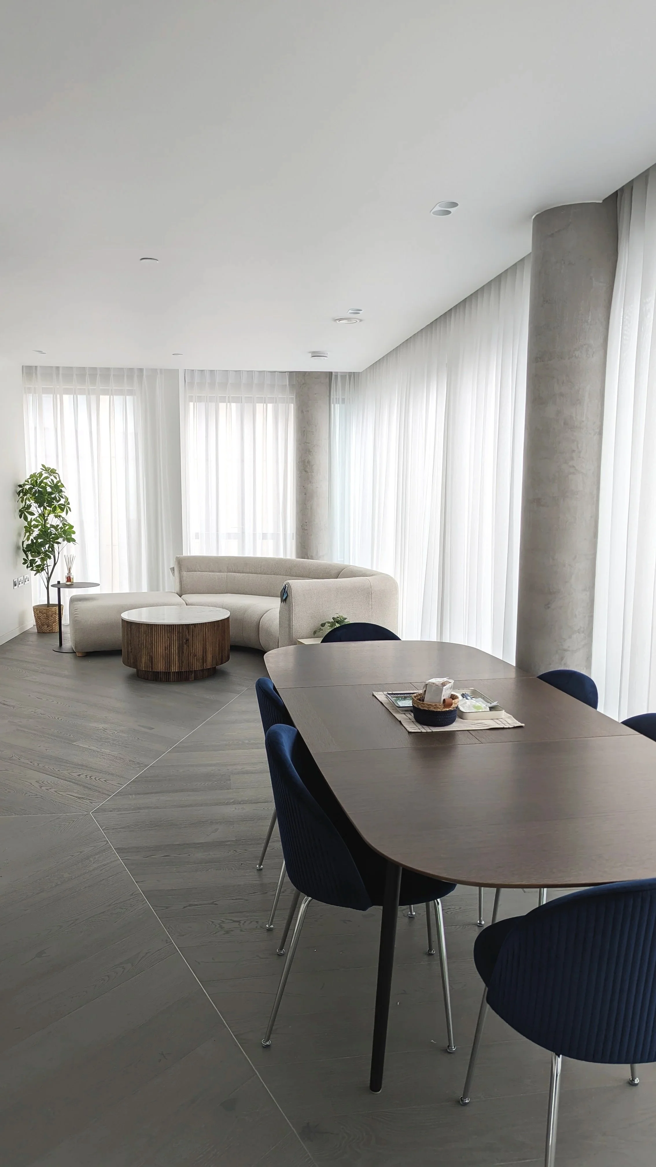 Modern living room with a curved white sofa, a round wooden coffee table, tall windows with sheer white curtains, a potted plant, and a dining table with blue chairs.