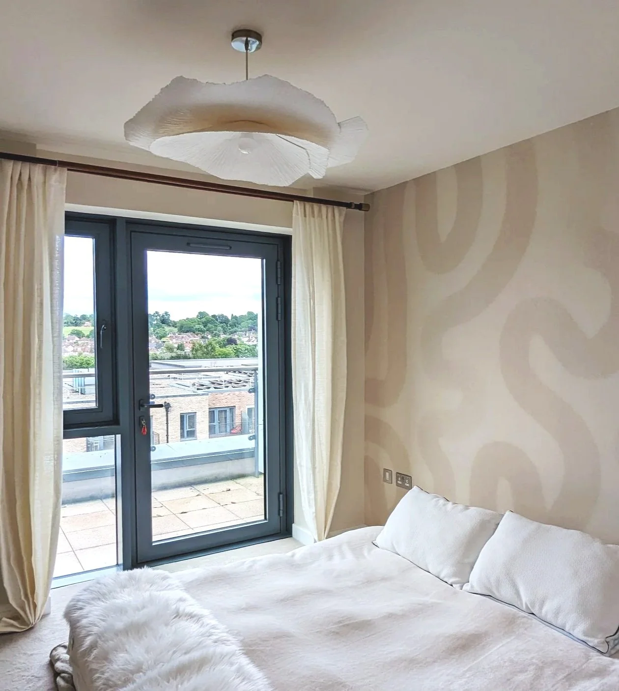 A bedroom with a bed covered in white bedding and pillows, open balcony glass door with cream curtains, and a view of a residential neighborhood with trees.