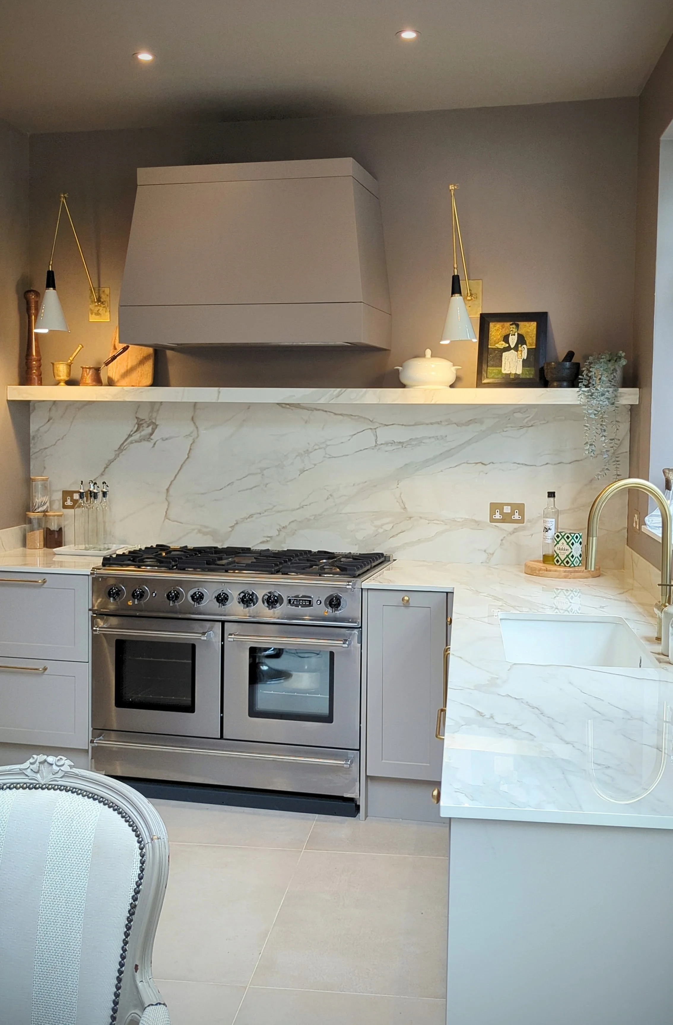 Modern kitchen with marble countertops, gray cabinets, and a stainless steel double oven. Decor includes wall-mounted lamps, framed artwork, plants, and kitchen accessories.