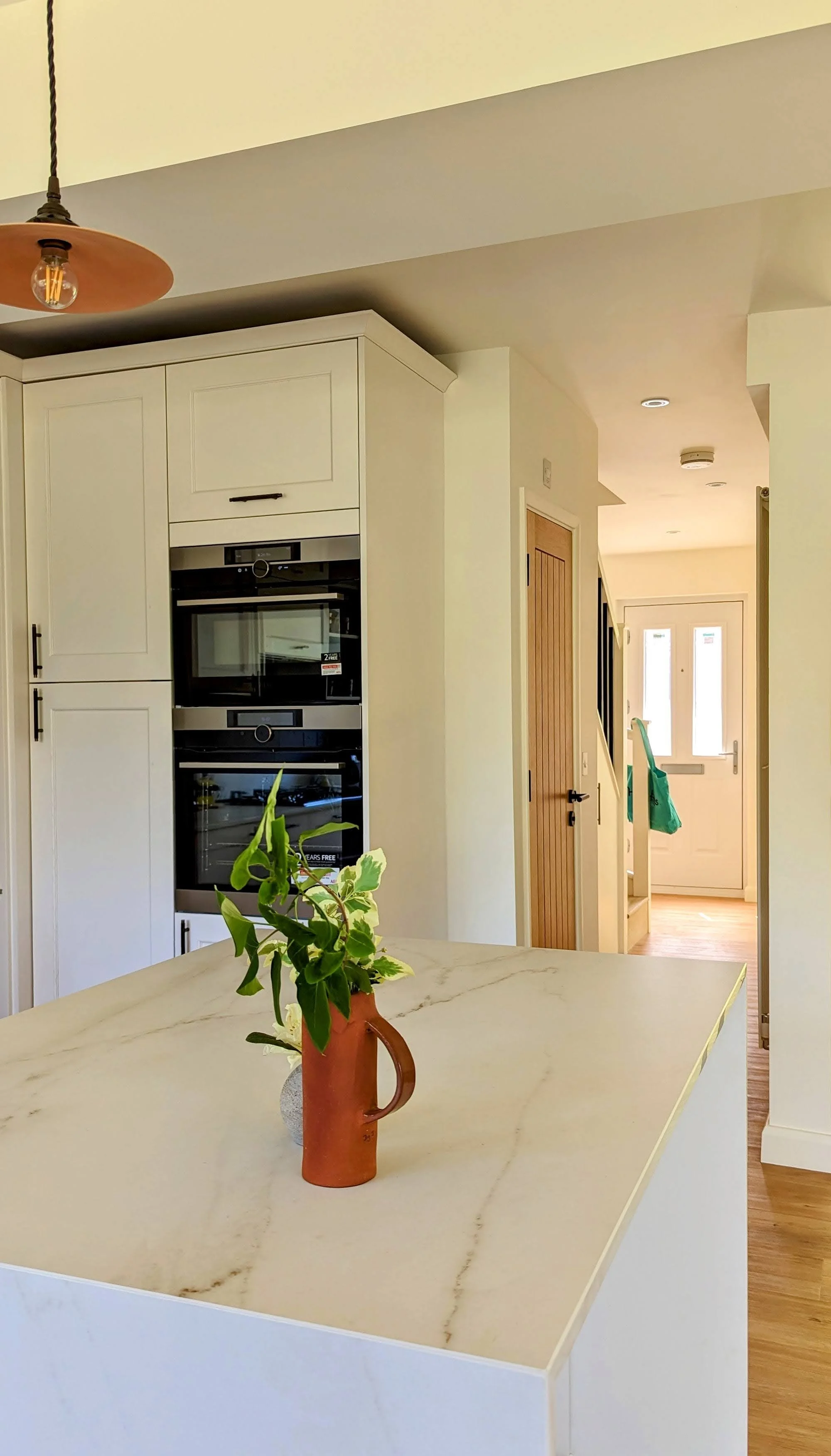 A kitchen with white cabinets, a built-in oven and microwave, and a marble countertop with a small plant in a brown pitcher. The background shows a hallway with a door and a window at the entrance.