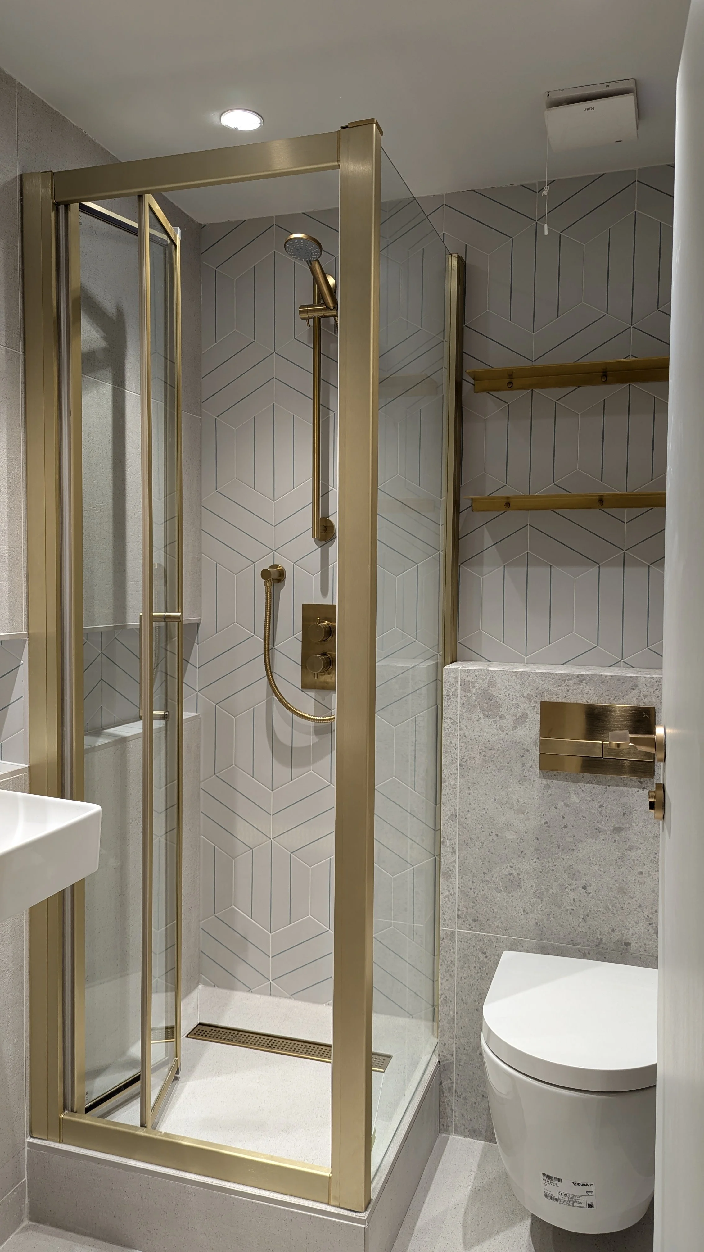 A modern bathroom with a brass fittings, wet room design with a small shower enclosure, hexagonal patterned wall tiles, brass metal shelf, and a sleek toilet.