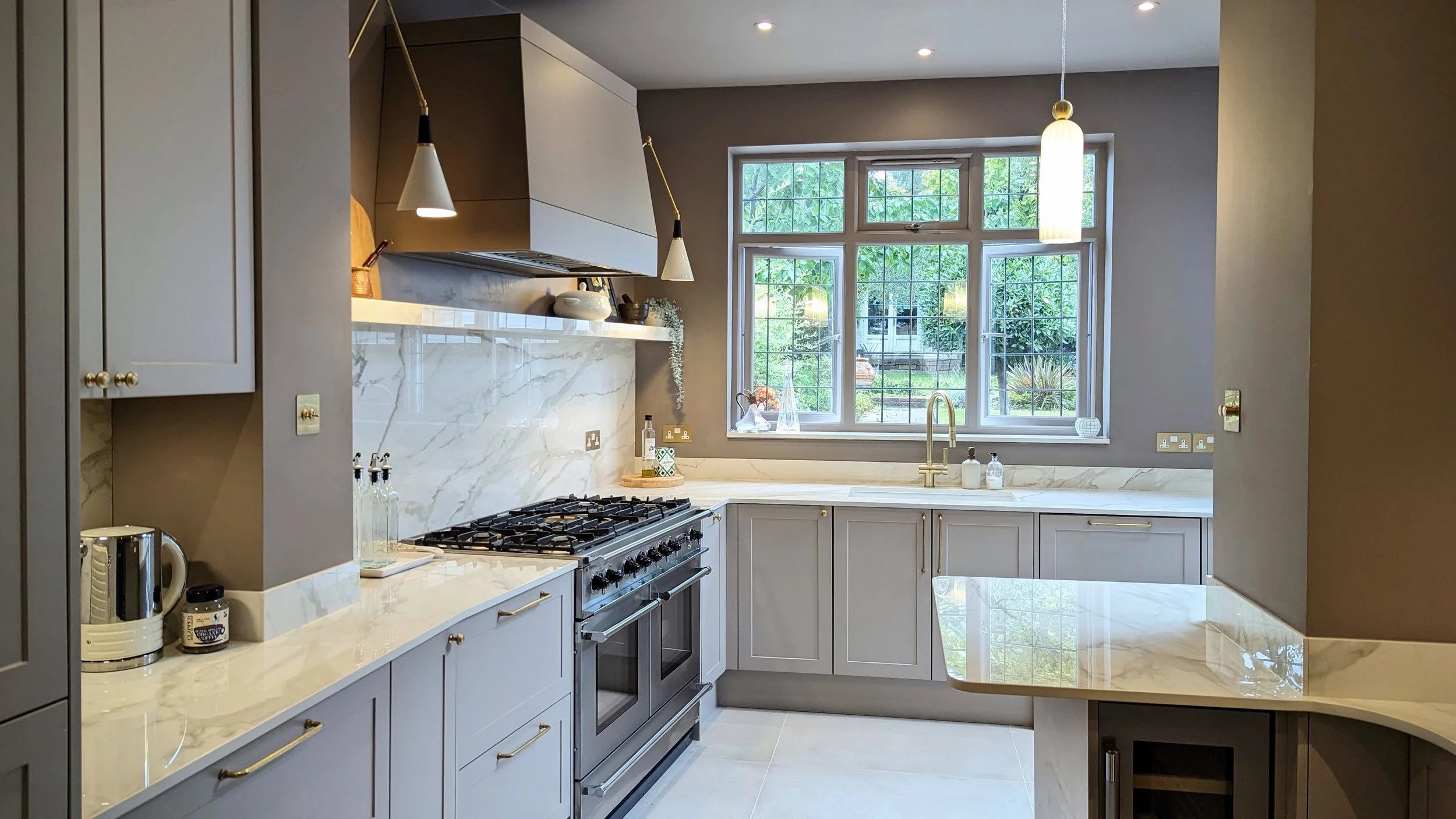 A modern kitchen with light gray cabinetry, a marble backsplash, and a large window overlooking greenery. The kitchen features a stainless steel stove, white marble countertops, and gold fixtures, including a faucet and cabinet knobs.
