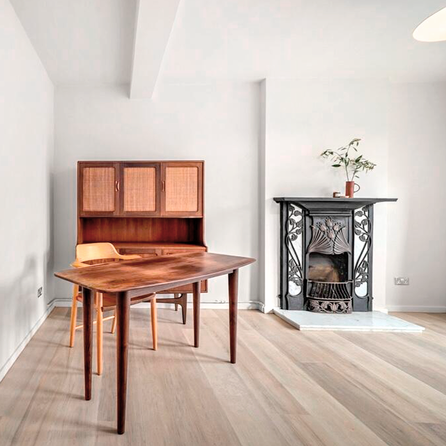 Minimalist living room with a mid century modern desk, and a popular make of a midcentury modern sideboard with rattan paneled doors. There is an Art Deco Style decorative fireplace with a plant on top, and a light-colored hardwood floor.