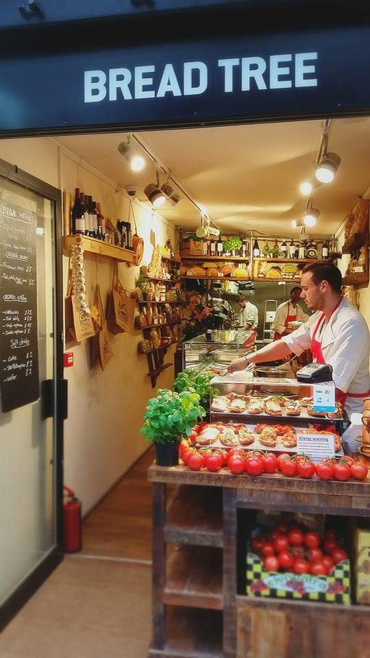 Inside boxpark,  'Bread Tree' is an italian eatery with staff preparing food behind a bespoke design wooden counter displaying tomatoes and baked goods, decorated with herbs and various bottles on shelves.