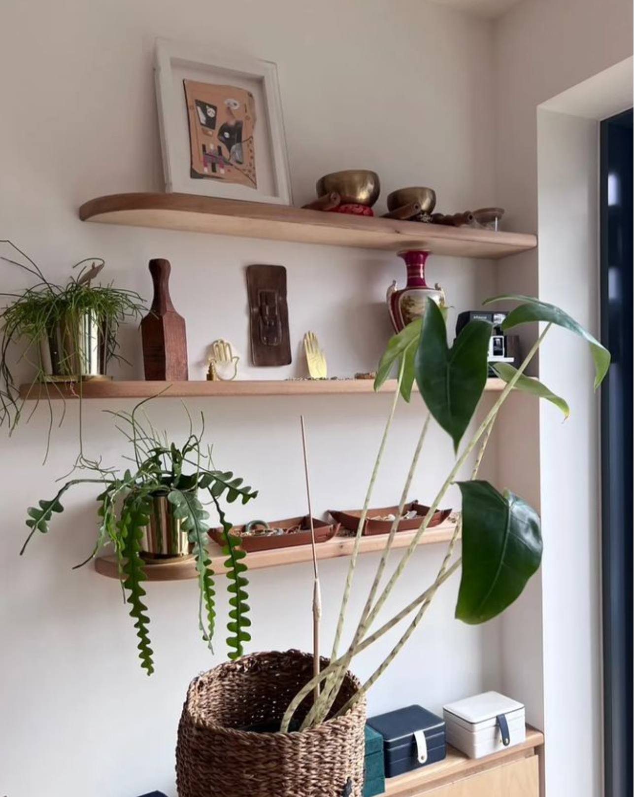 Interior wall with three wooden custom made shelves displaying decorative items, including framed artwork, brass cups, a vase, and small sculptures and plants, with a wicker basket and storage boxes below.