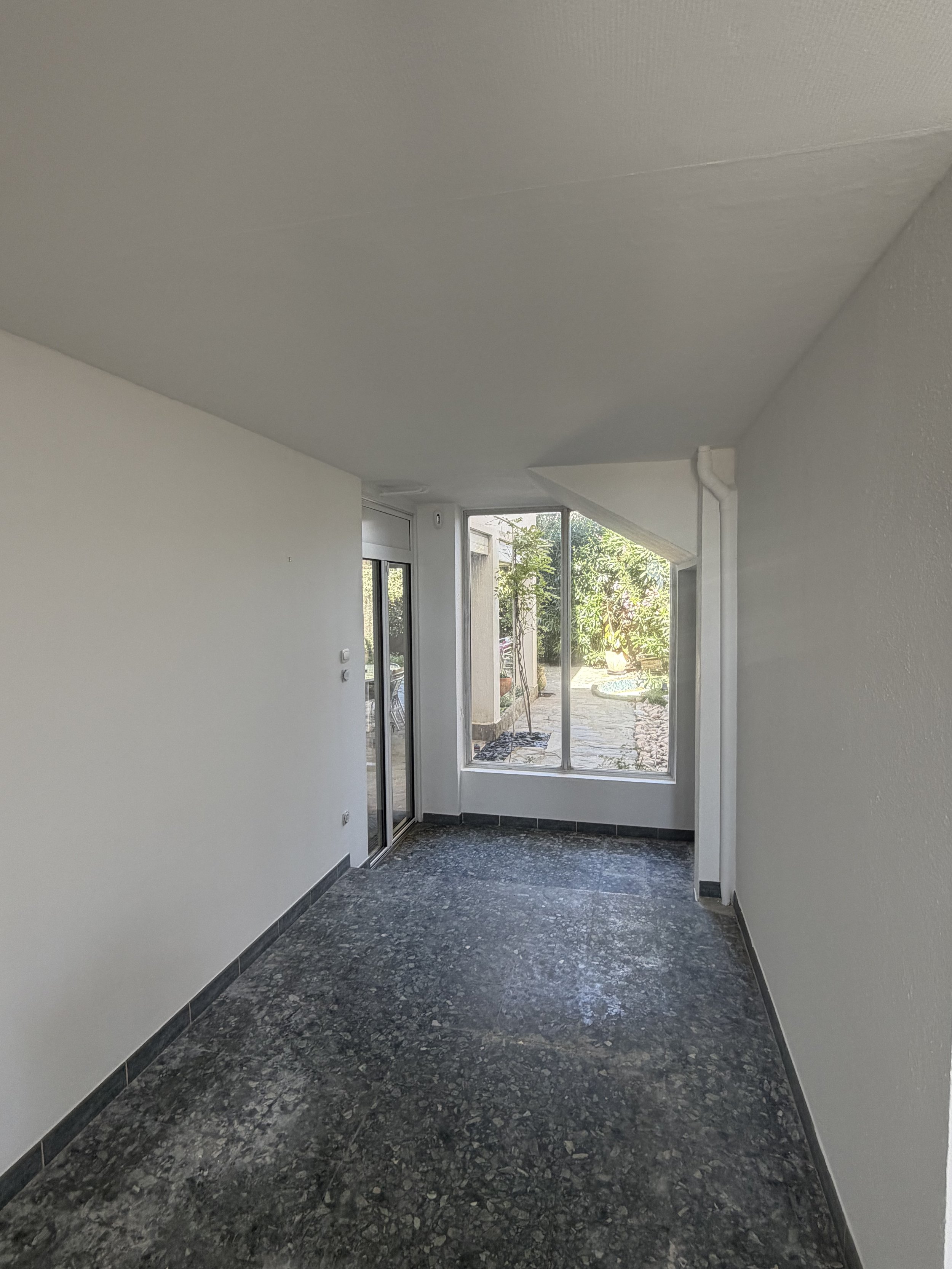 Intérieur d'une pièce lumineuse avec murs blancs, sol en pierre noire et grande fenêtre donnant sur un jardin extérieur.
