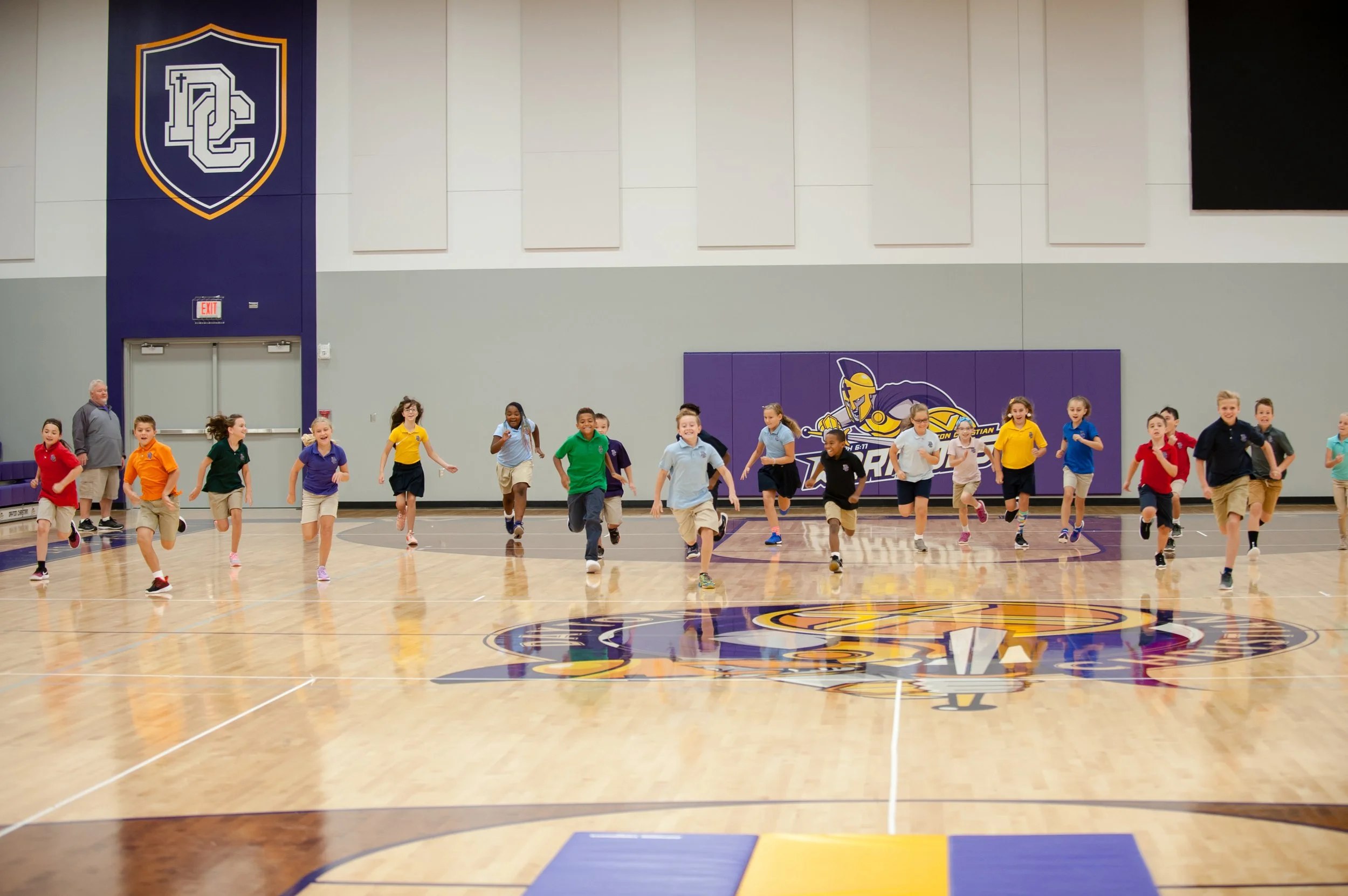 A group of children running in a gymnasium with a basketball court and sports logos on the wall.