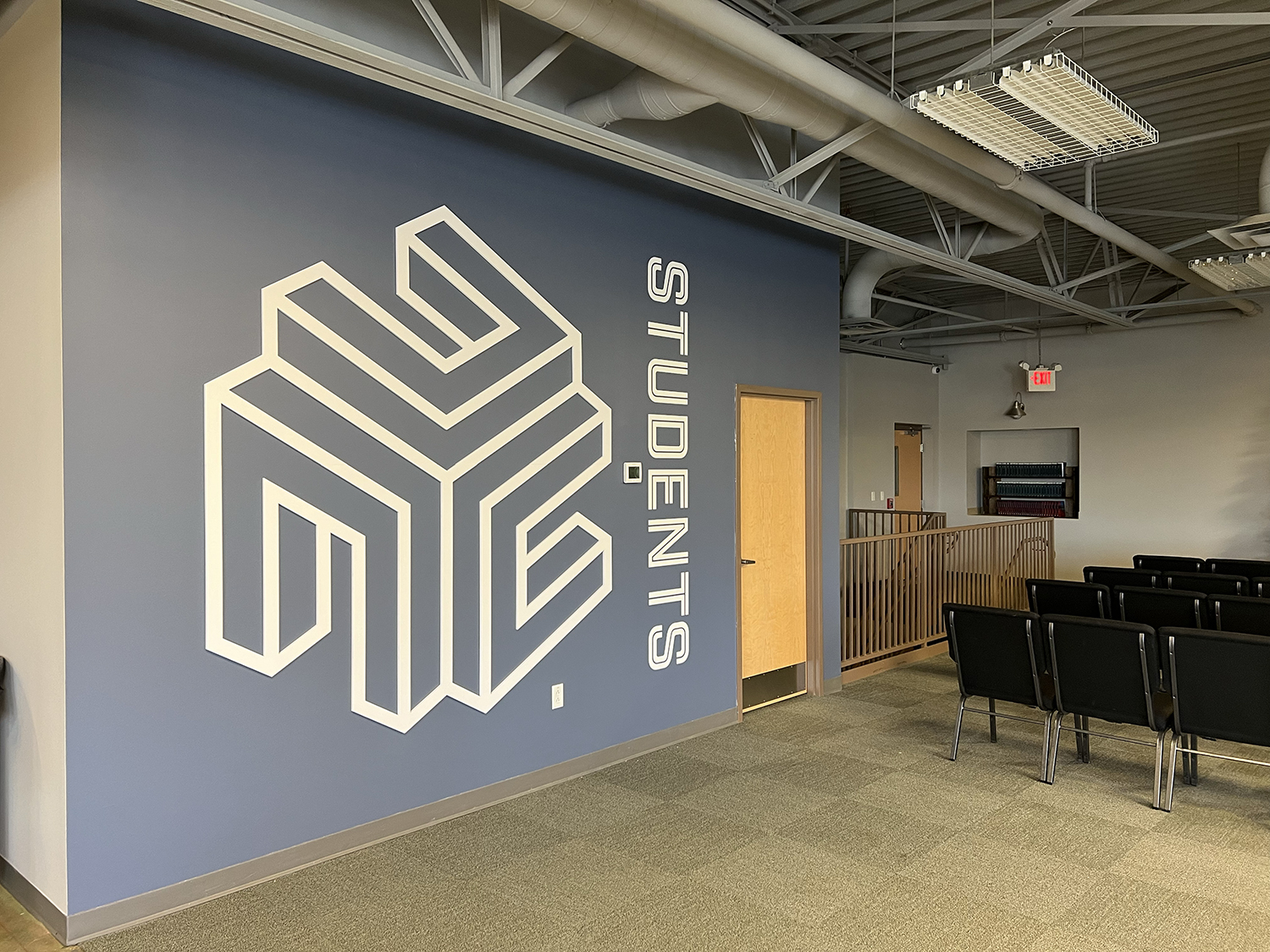 Interior of a room with a large wall decal reading 'STUDENTS' and a geometric logo, with rows of black chairs, a wooden door, and an exit sign visible.