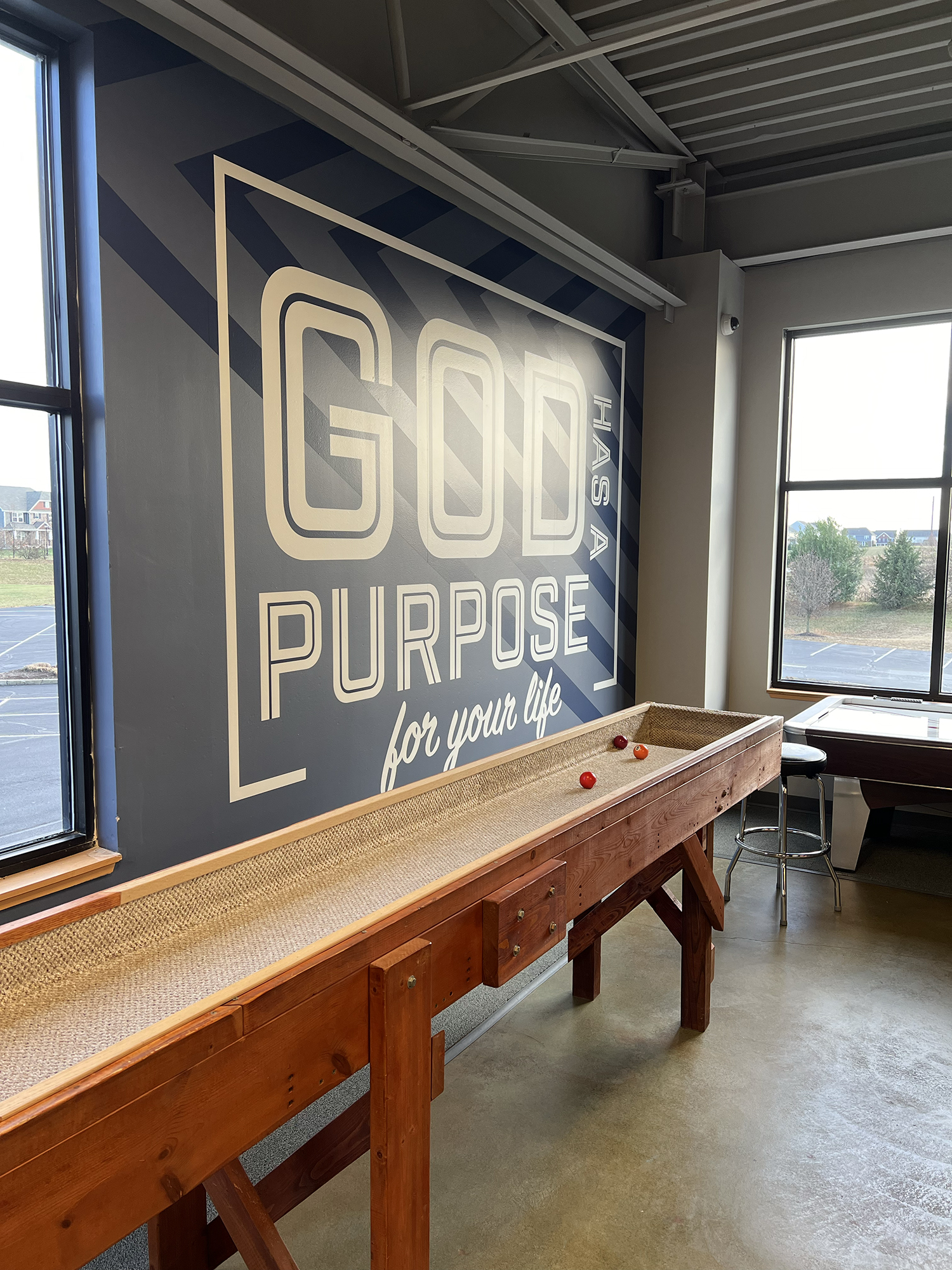 An indoor space with a large wall mural that says "GOD PURPOSE" and "for your life". A wooden shuffleboard table with pucks on it is in the foreground. There are windows on the left and right sides, letting in natural light.
