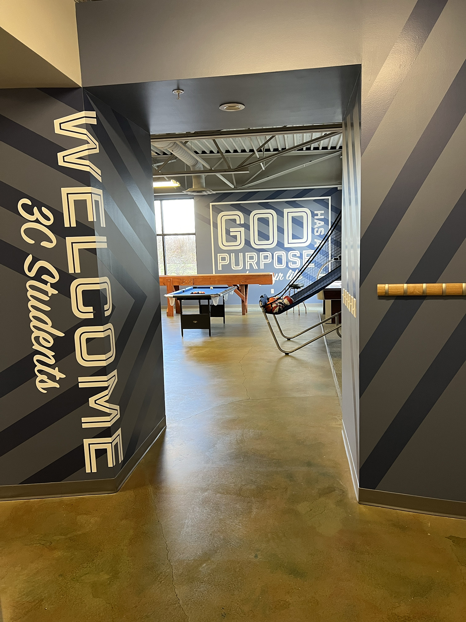 Interior view of a recreational space featuring a pool table, signs with motivational phrases, and a unique swing with a helmet on it.
