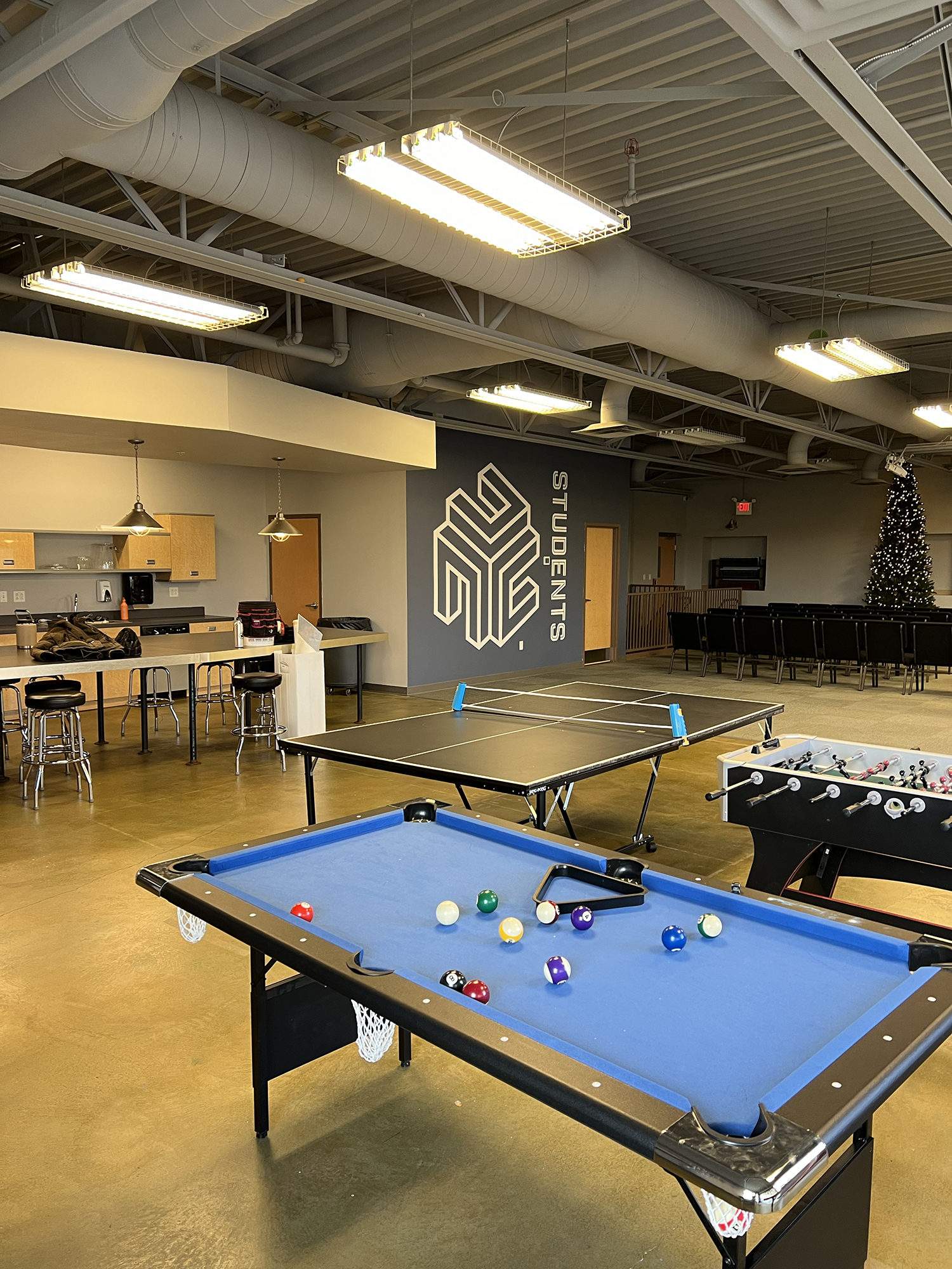 Pool hall with blue billiards table and pool balls, foosball table, ping pong table, chairs, Christmas tree, and study studies sign on the wall.