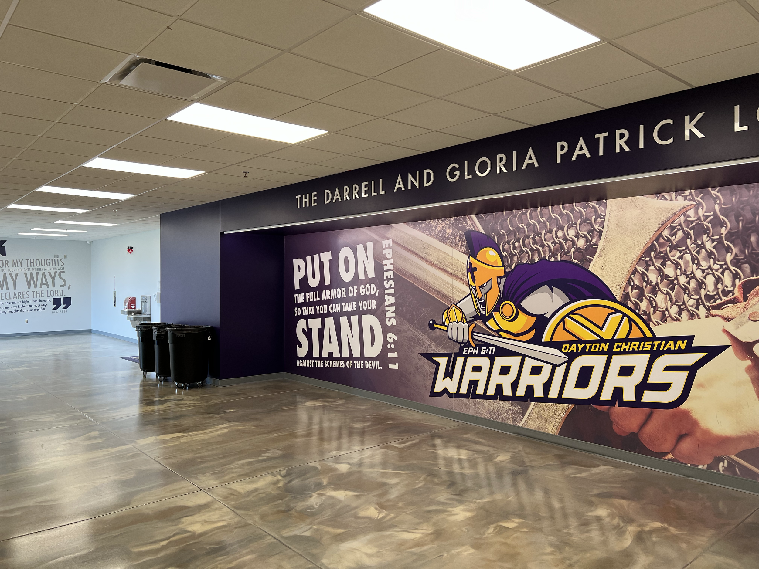 Indoor hallway with beige ceiling tiles and fluorescent lights. There is a black wall with a large colorful poster promoting Dayton Christian Warriors, featuring a cartoon knight with a sword and shield. Part of a white wall with motivational text an