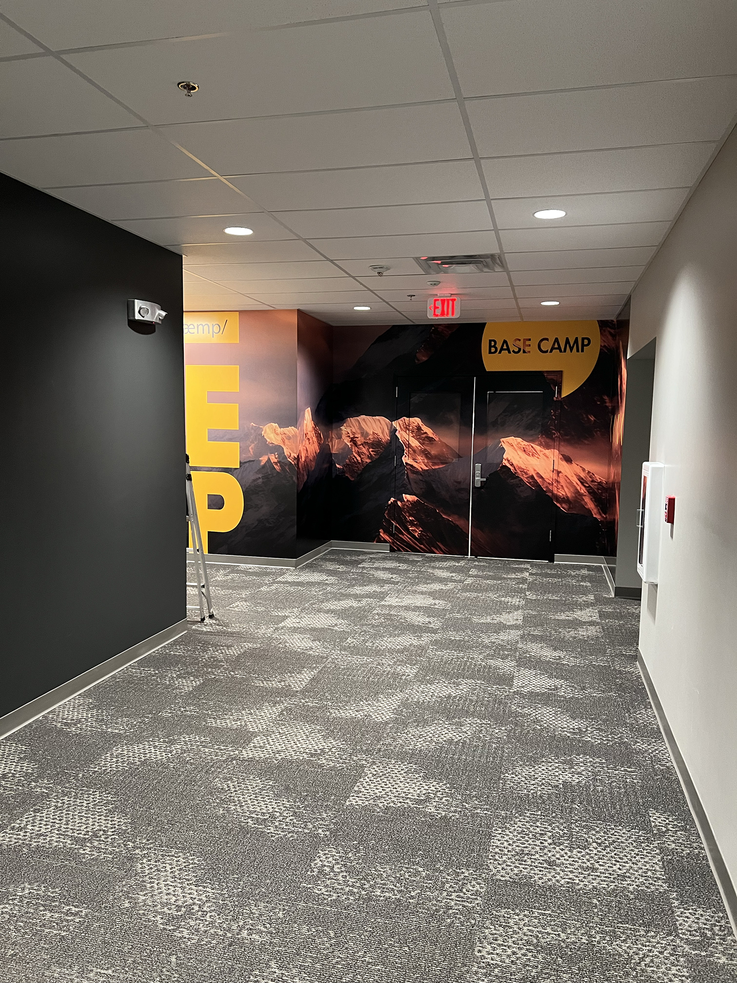 An indoor hallway with gray and black walls, gray carpeted floor, and ceiling tiles. There is a security camera on one black wall, an emergency exit door with a red illuminated 'EXIT' sign, and a mural of mountains on the back wall with the text 'BAS