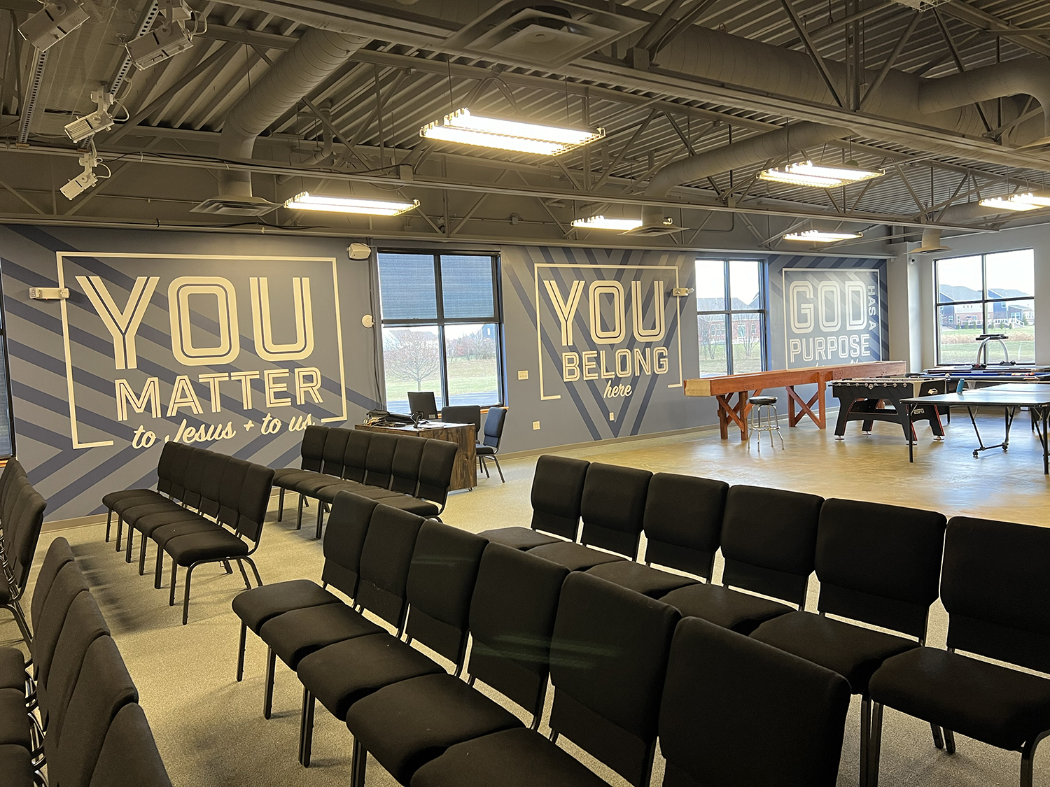 An indoor space with rows of black chairs, motivational wall murals with phrases like "You matter to Jesus + to us" and "You belong here," large windows, and recreational tables like ping pong and foosball.