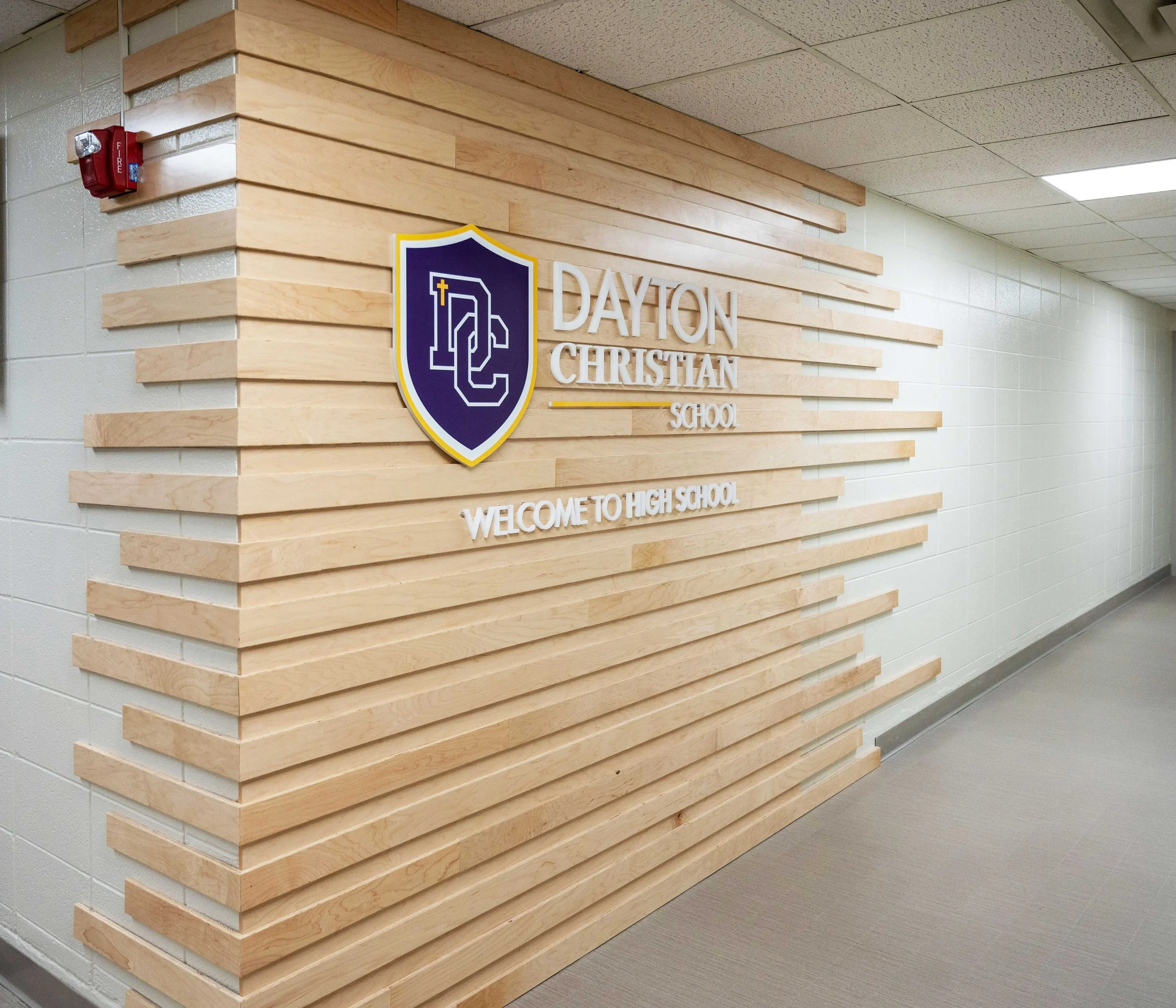 Interior of a school hallway with a wooden paneled wall displaying the Dayton Christian School logo and name. The logo includes a shield with a cross and a stylized letter 'D'. There is a fire alarm mounted on the wall near the corner. The ceiling ha