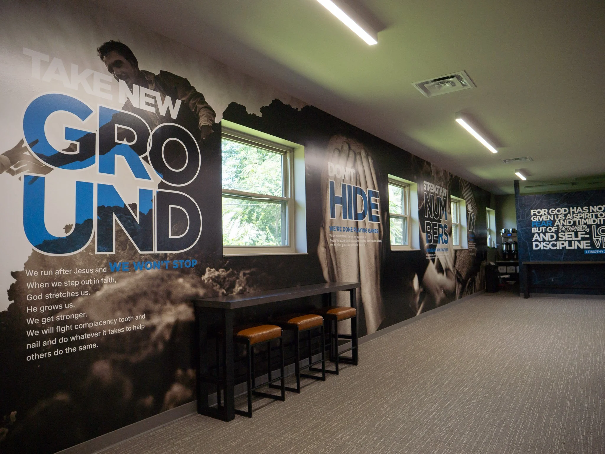 Indoor room with motivational posters on the wall, featuring phrases like "TAKE NEW GROUND," "DON'T HIDE," and others. There are large windows letting in natural light, and a row of chairs with brown seats along the wall. The ceiling has modern light