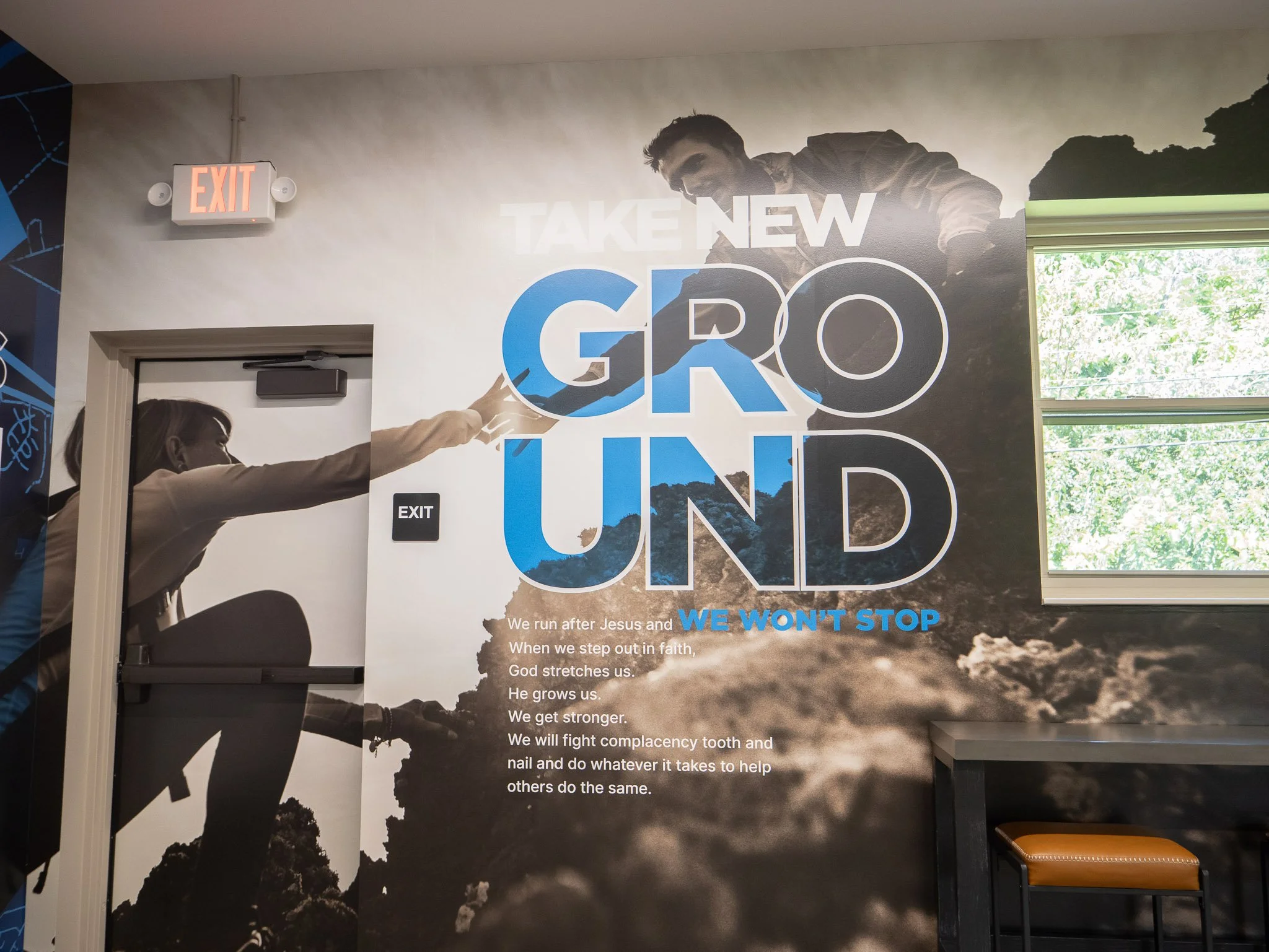 Motivational church-like poster with an image of a man reaching out to a person, text that reads 'TAKE NEW GROUND' and a Bible verse about faith and perseverance, near an exit sign and window showing green trees outside.