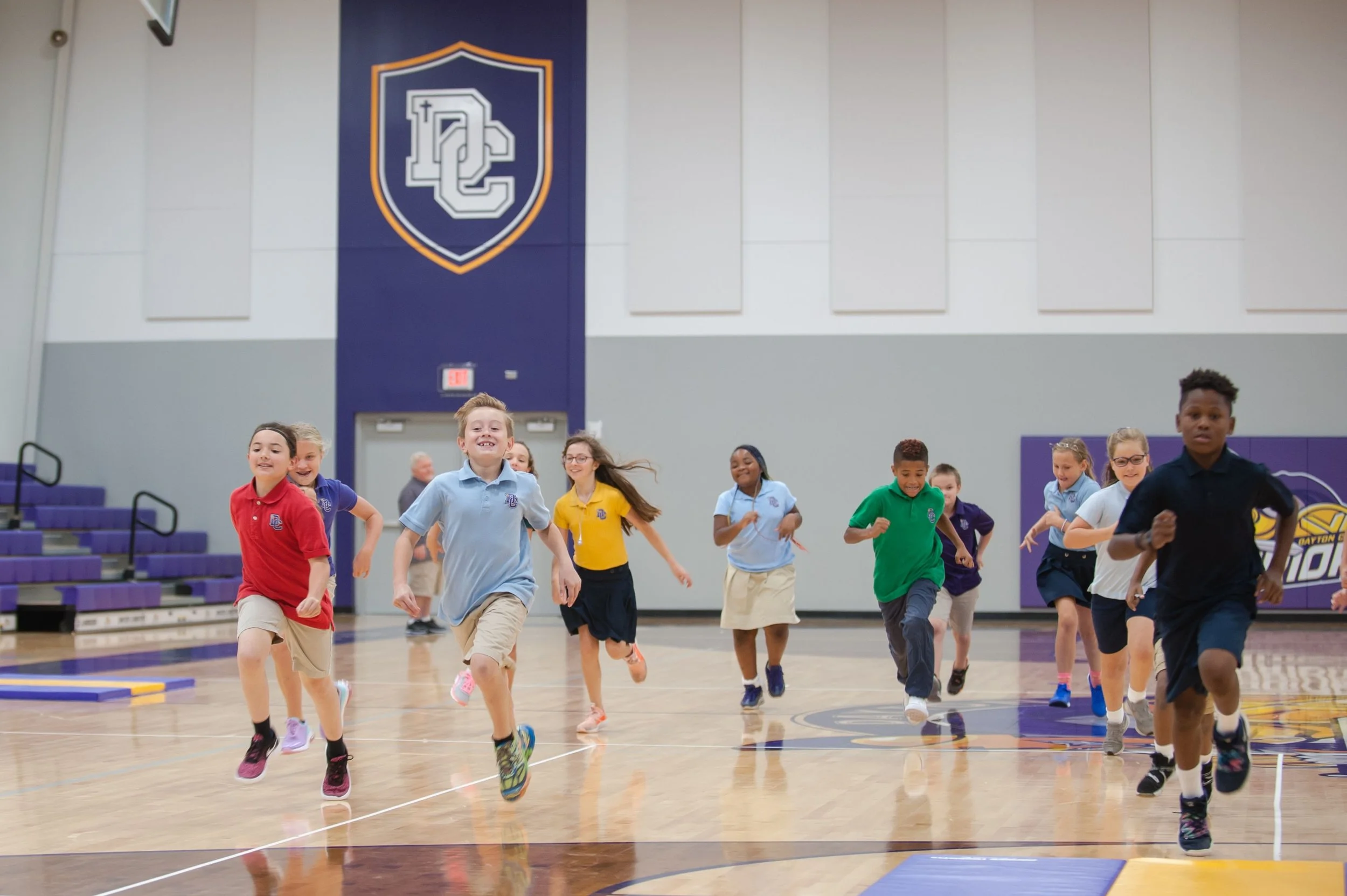 Children running in a gymnasium with a large school logo on the wall.