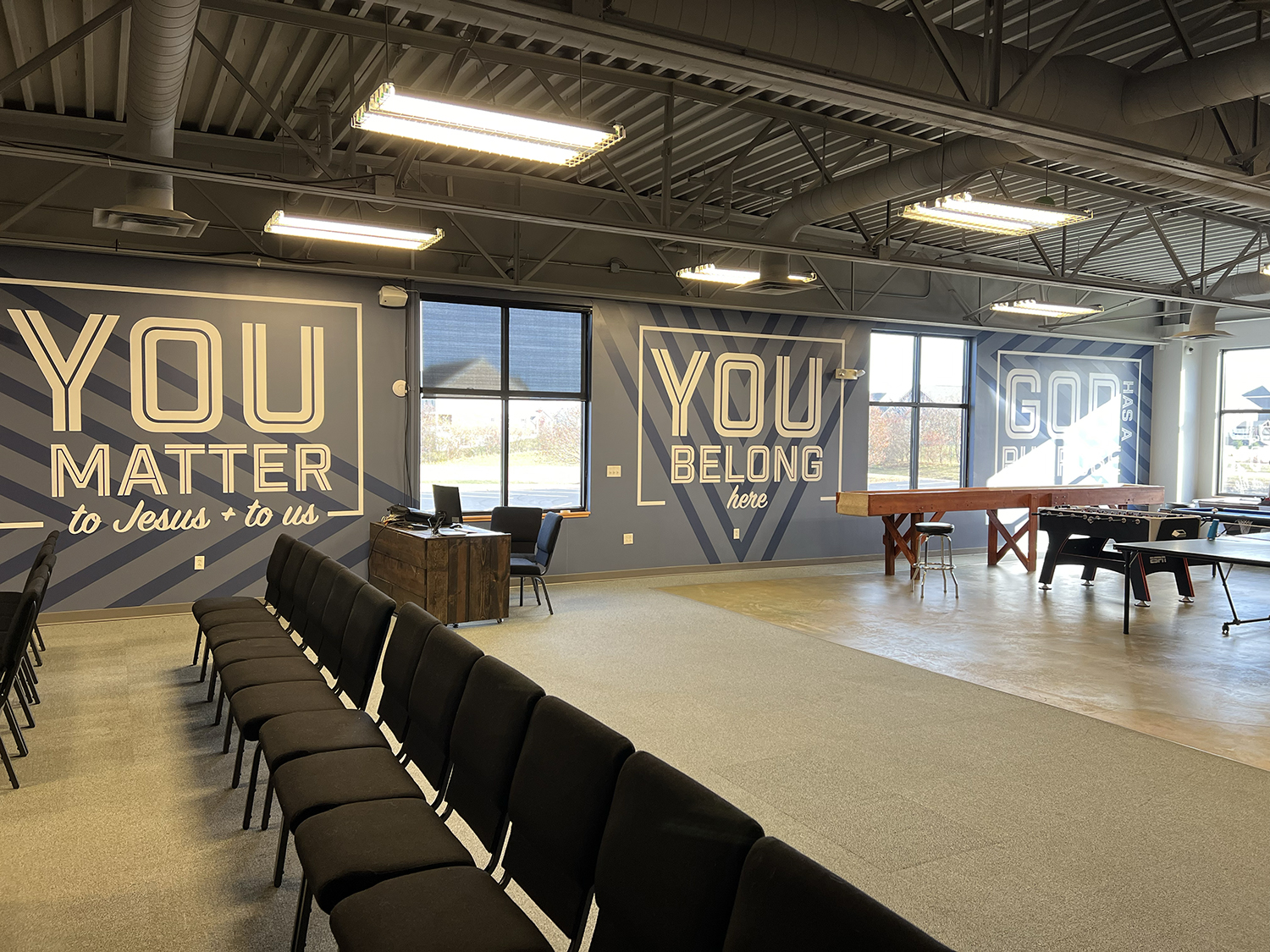 A spacious room with large windows and motivational church-themed wall murals that read "You matter to Jesus + to us," "You belong here," and "God has a plan for you." The room is set up with black chairs in the foreground, a wooden desk with a compu