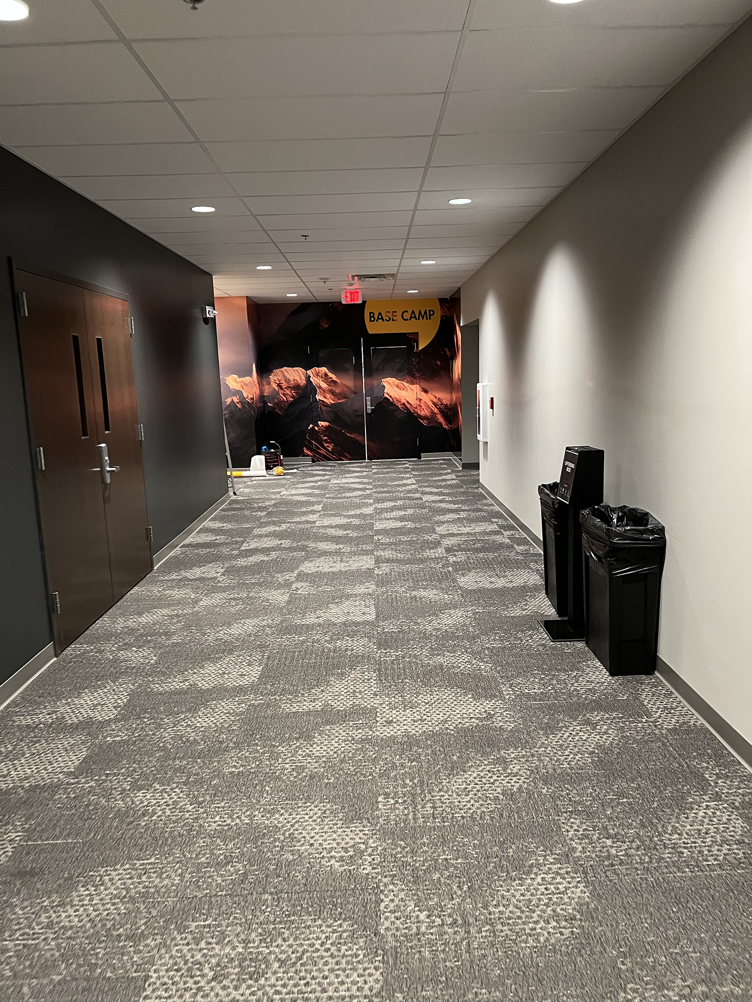 A hallway in a building with patterned carpet, white and black walls, a mural of mountains on the far end, and a sign that reads 'Base Camp' hanging above the mountains mural.