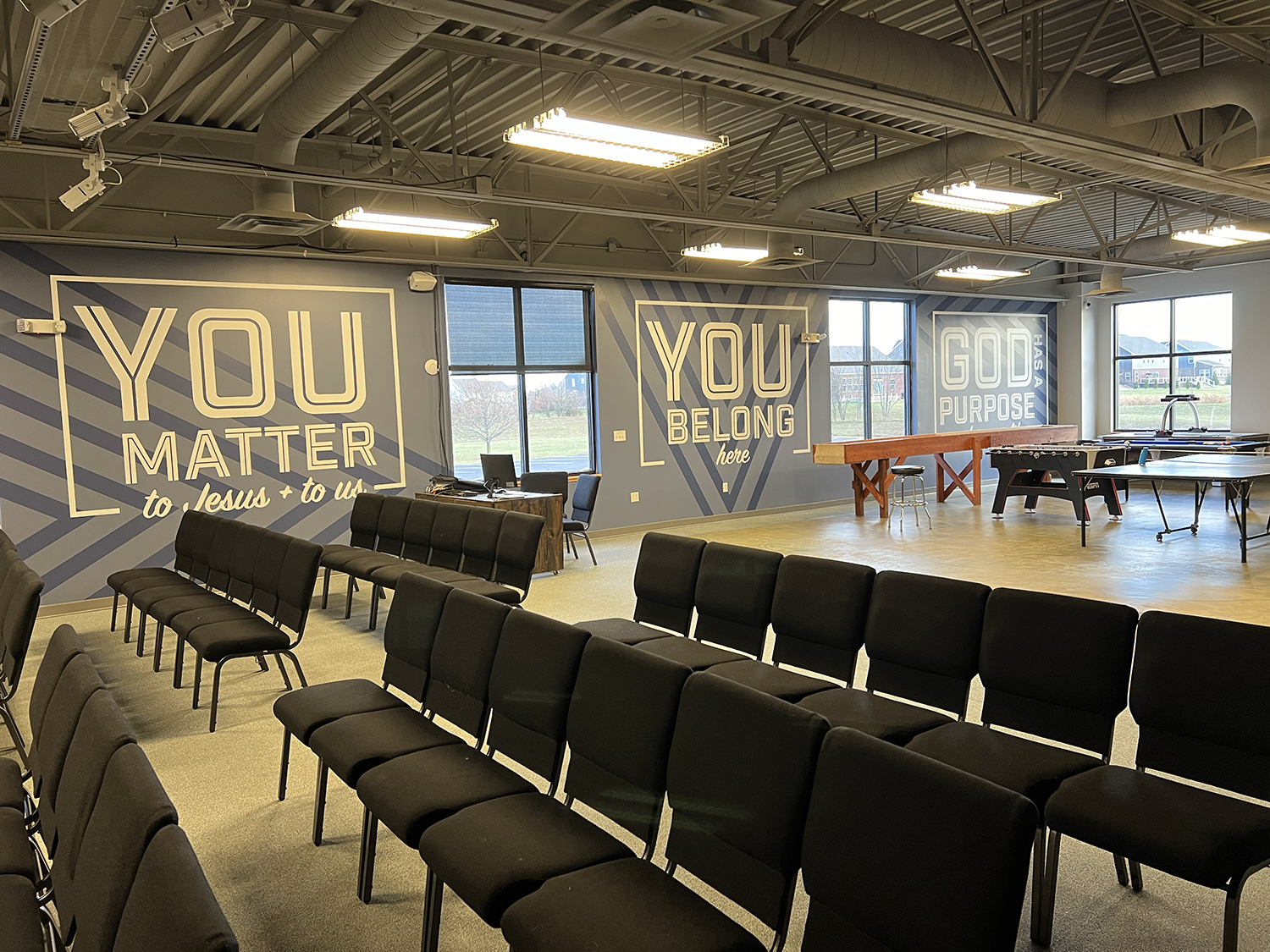 Interior of a church or meeting space with rows of black chairs, painted wall with motivational messages, windows showing a grassy landscape outside, and exercise equipment including a ping pong table and foosball table.