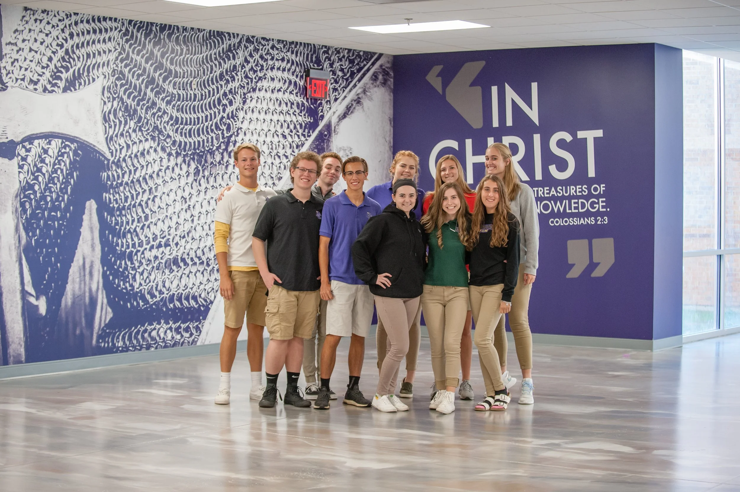 Group of nine young people, smiling, posing in front of a large wall with an abstract design and a sign that says 'IN CHRIST' and a Bible verse reference 'Colossians 2:3'.