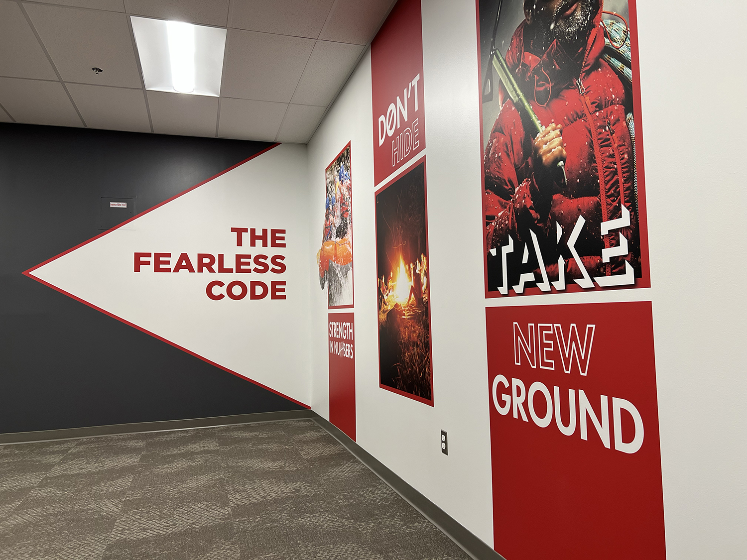 A hallway with wall artwork and text promoting a fearless code, with phrases such as "The Fearless Code," "Don't Hide," "Take," and "New Ground." The wall has images of outdoors and adventure themes, including a person in a red jacket and a fire scen