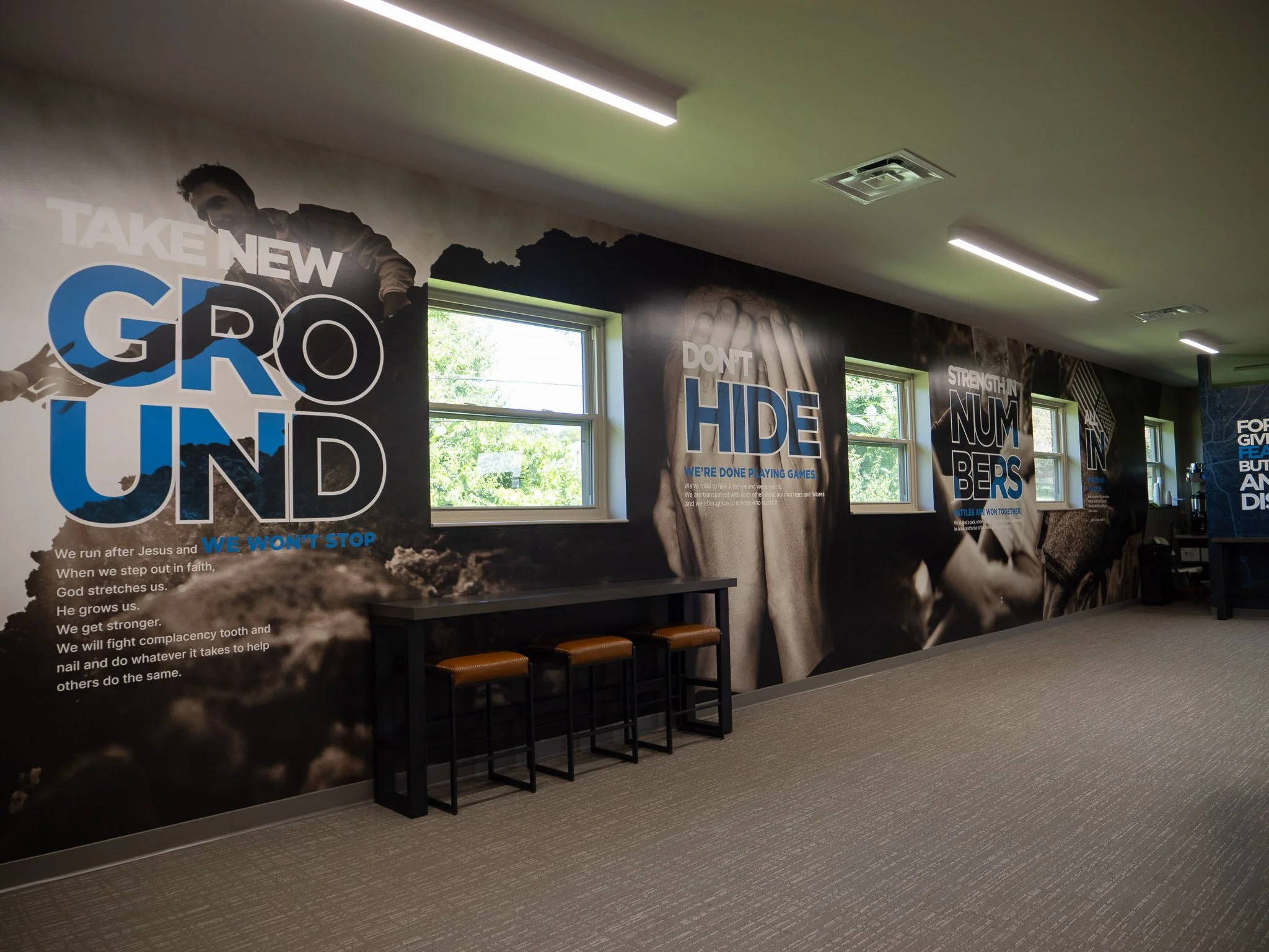 A wall with inspirational Fearless code posters, including phrases like 'Take new ground', 'Don't hide', and 'Strength in numbers', located in a room with windows and chairs beneath the posters.