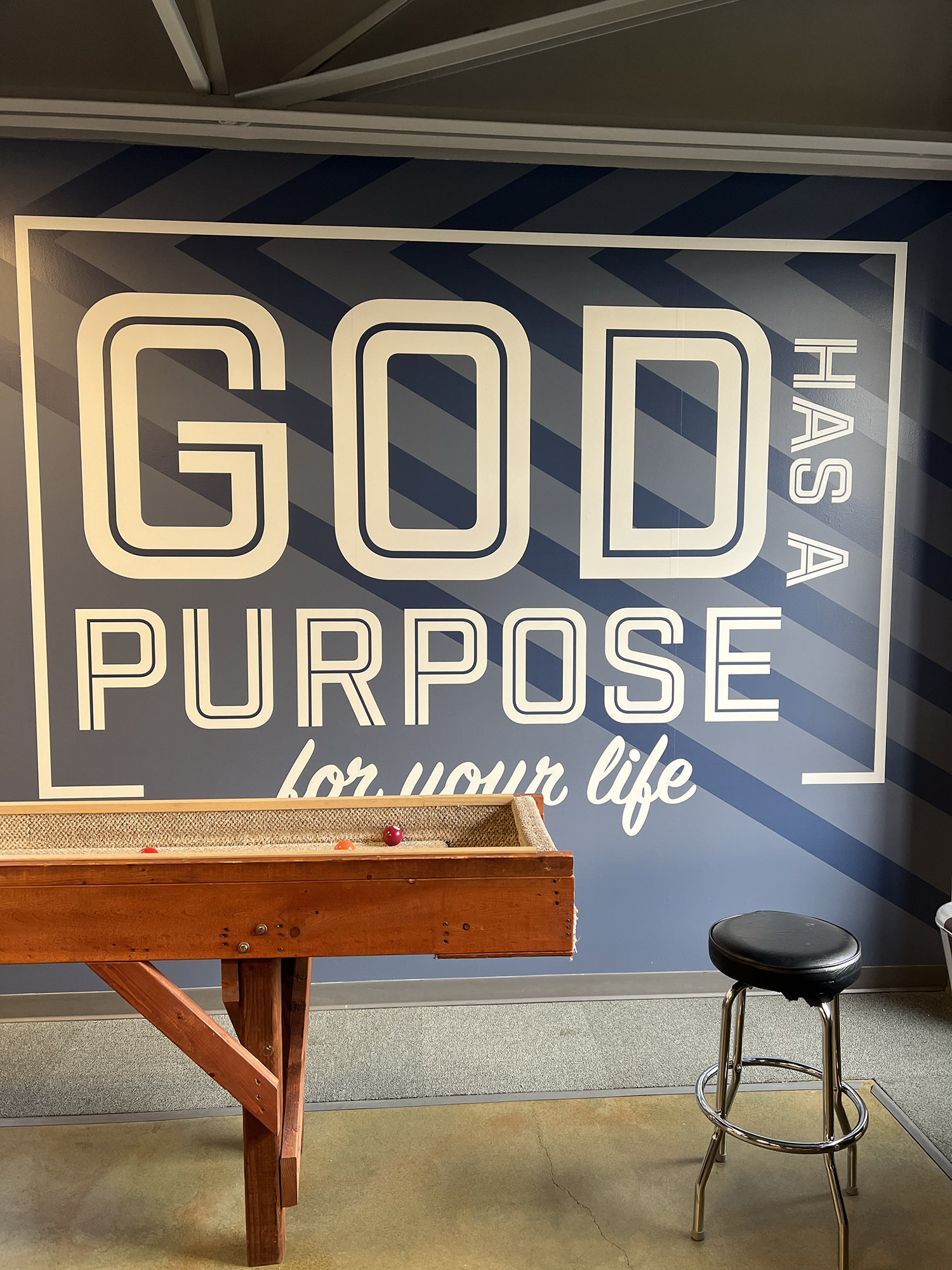 A wooden billiards table with balls on it, set against a wall with a mural that reads 'GOD PURPOSE for your life' in large white letters and dark diagonal stripes in the background. There is a stool with a black cushioned seat to the right of the tab