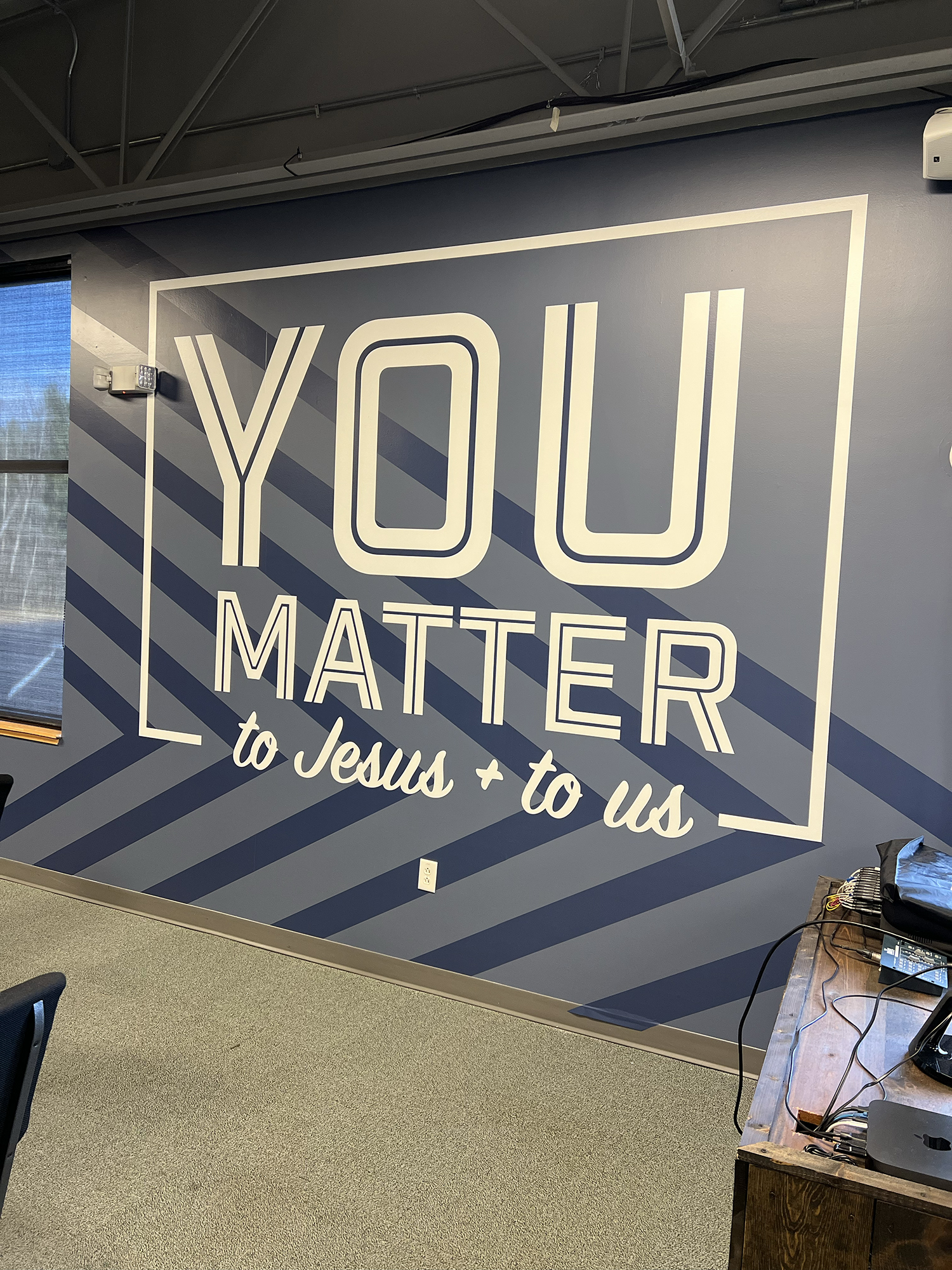 A large wall decal with the words "YLOU MATTER" and a partially visible phrase "to Jesus + to us" underneath, in an indoor space with a gray carpeted floor and a wooden table with electronic equipment on it.