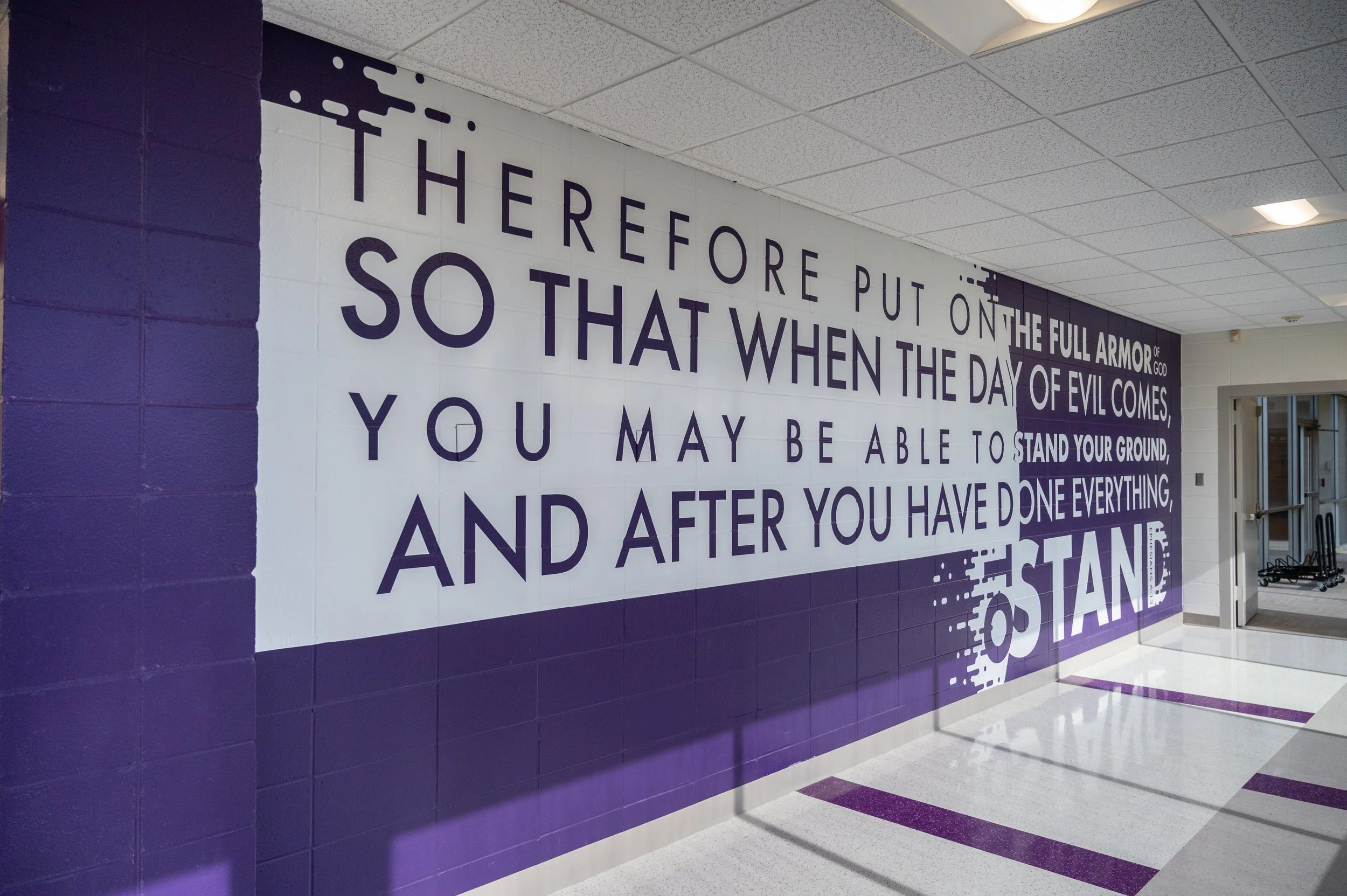 Purple and white wall with quote in purple lettering, part of a school or gym interior, with visible ceiling tiles and part of a doorway.