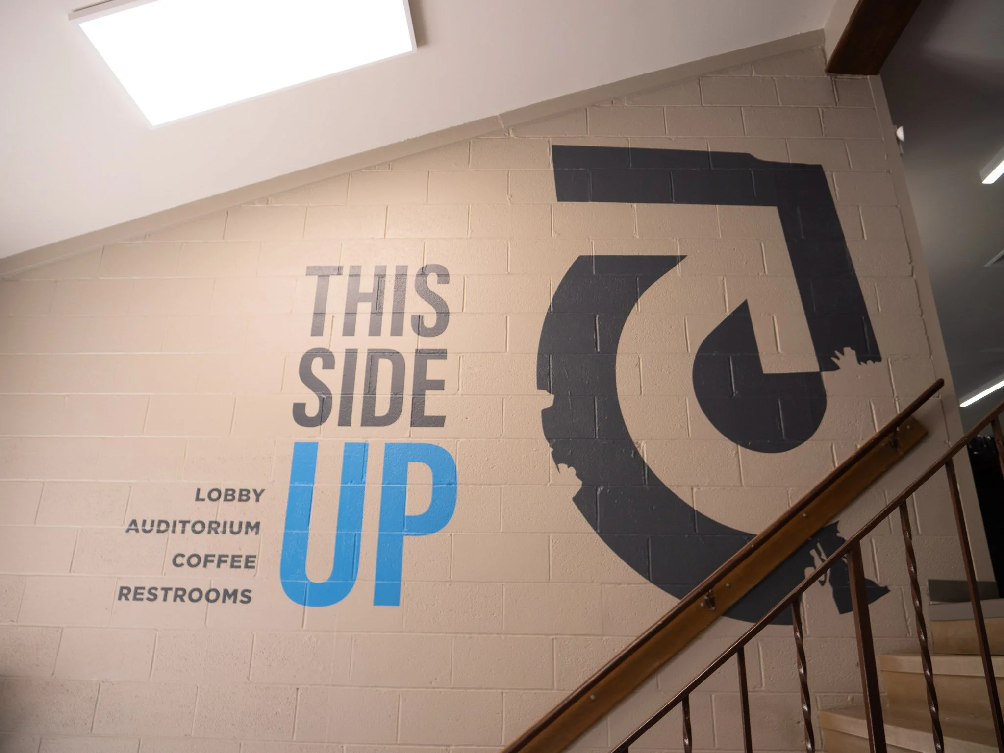 Sign on a wall with blue and black text indicating directions to various rooms, including lobby, auditorium, coffee, and restrooms. The sign features a large black arrow pointing right and the phrase "This Side Up" painted on the wall.
