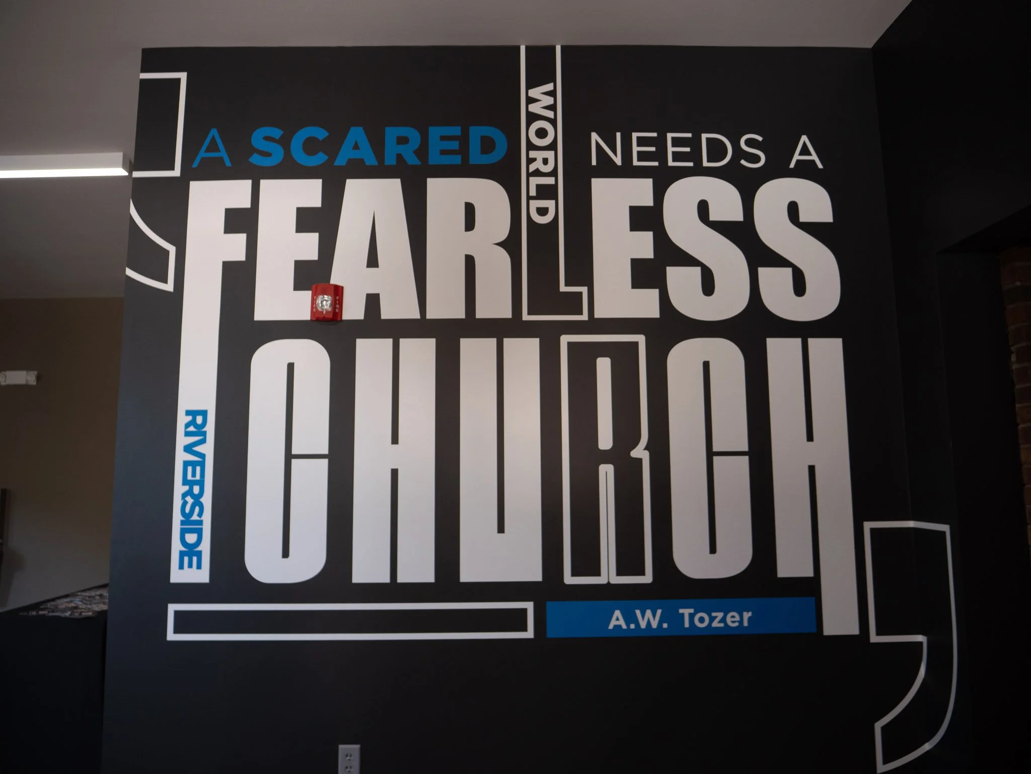 Wall display with large, bold white and blue text reading "A SCARED WORLD NEEDS A FEARLESS CHURCH" and "A.W. Tozer" on a black background, with some decorative white lines and shapes.