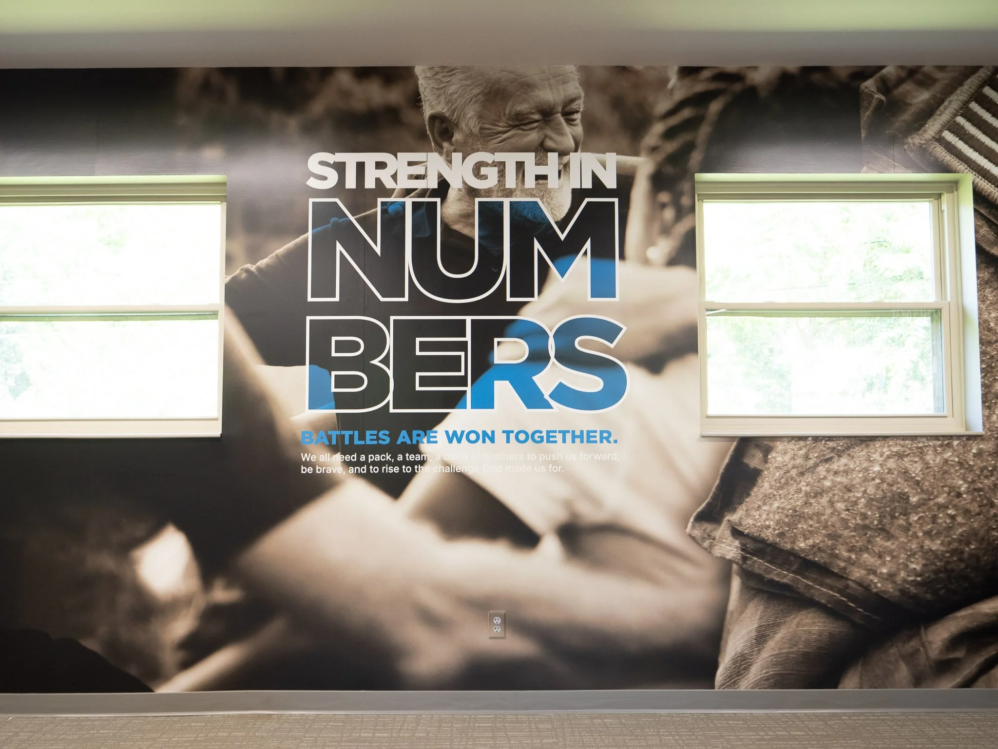 A large wall poster featuring an elderly man with a joyful expression, the text "Strength in Numbers" and "BATTLES ARE WON TOGETHER," along with a message about teamwork and perseverance.
