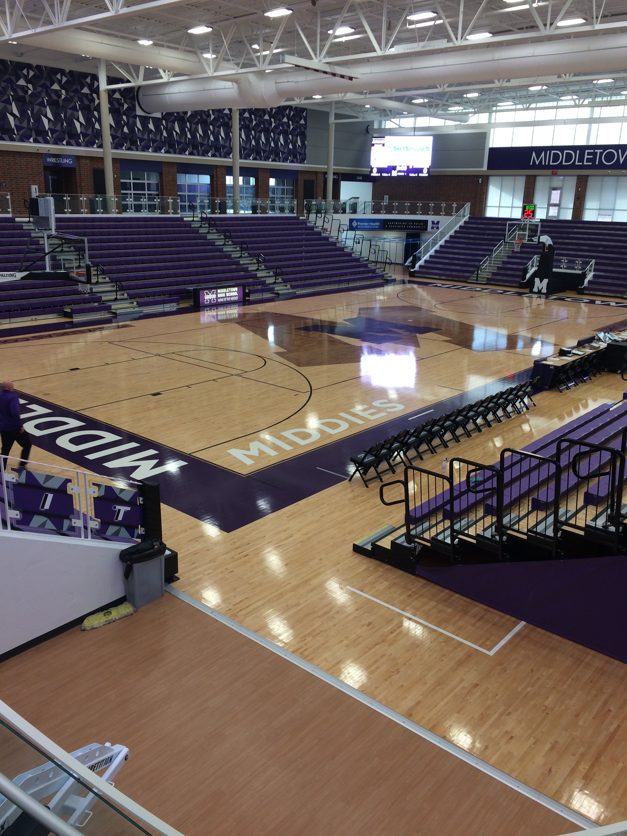 Empty indoor basketball court with purple and gold seating, digital scoreboard, and large windows at Middleton High School gym.