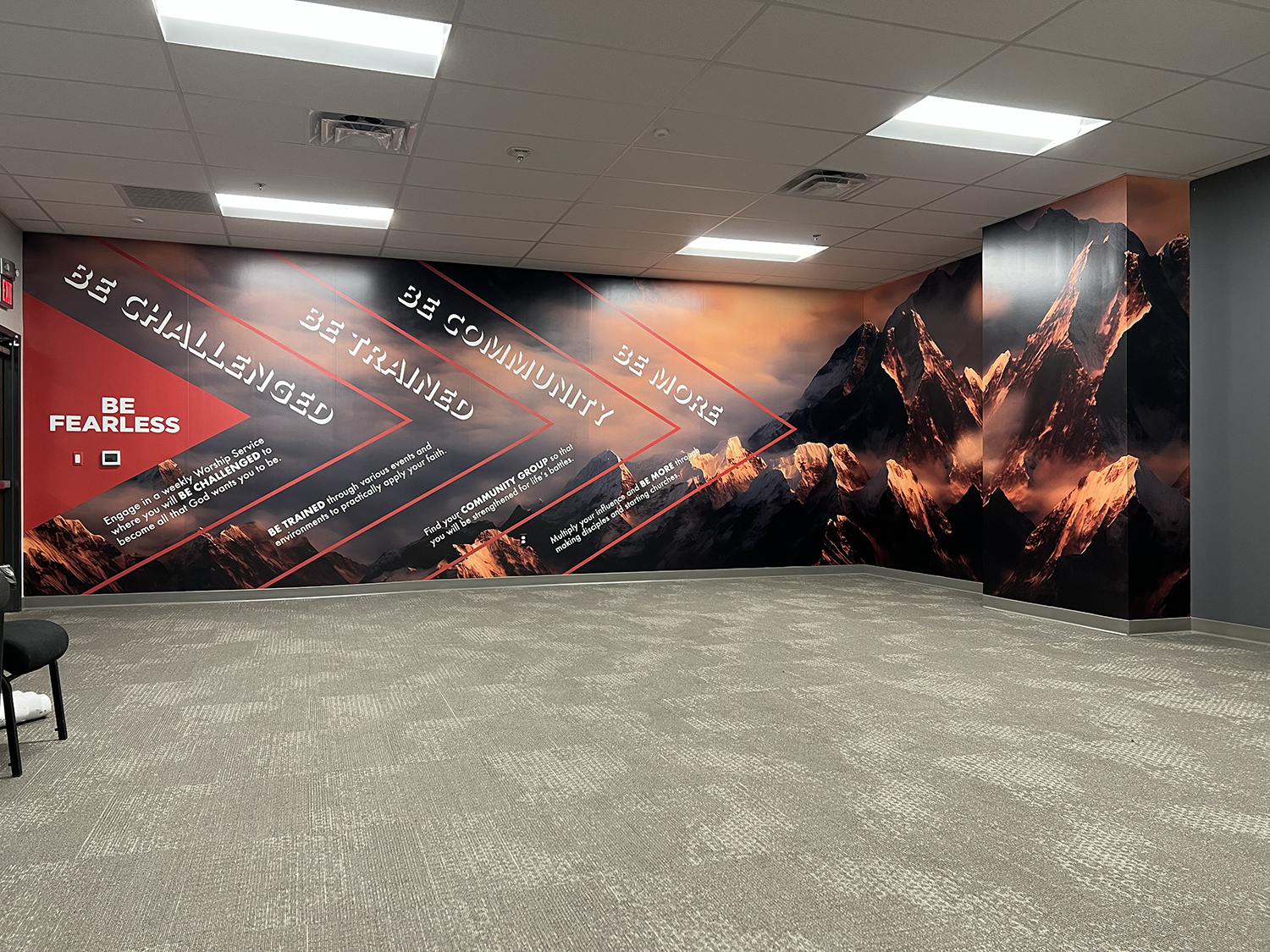 Interior of a room with a large mural on the wall depicting snow-capped mountains and a sunset sky. The mural contains motivational text about community, faith, and courage.