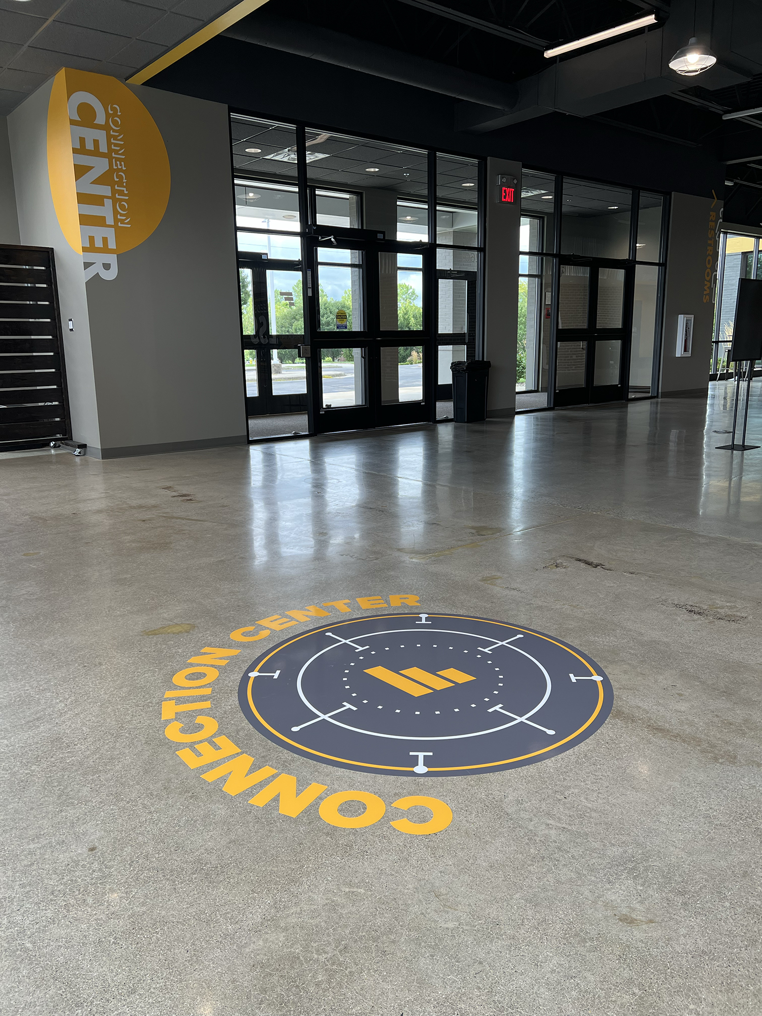 Interior of a building with glass doors, a yellow and black emblem on the floor reading 'CONNECTION CENTER,' a yellow circular sign on the wall reading 'CENTER RETIRING,' emergency exit sign, and bright outdoor scenery visible through the doors.