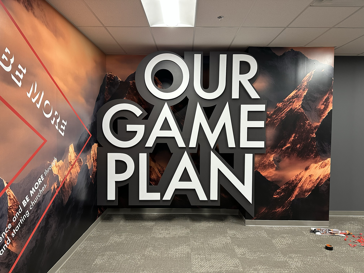 Interior wall with large 3D letters spelling 'OUR GAME PLAN' against a mountain landscape background, and part of a red outline with text about church outreach.
