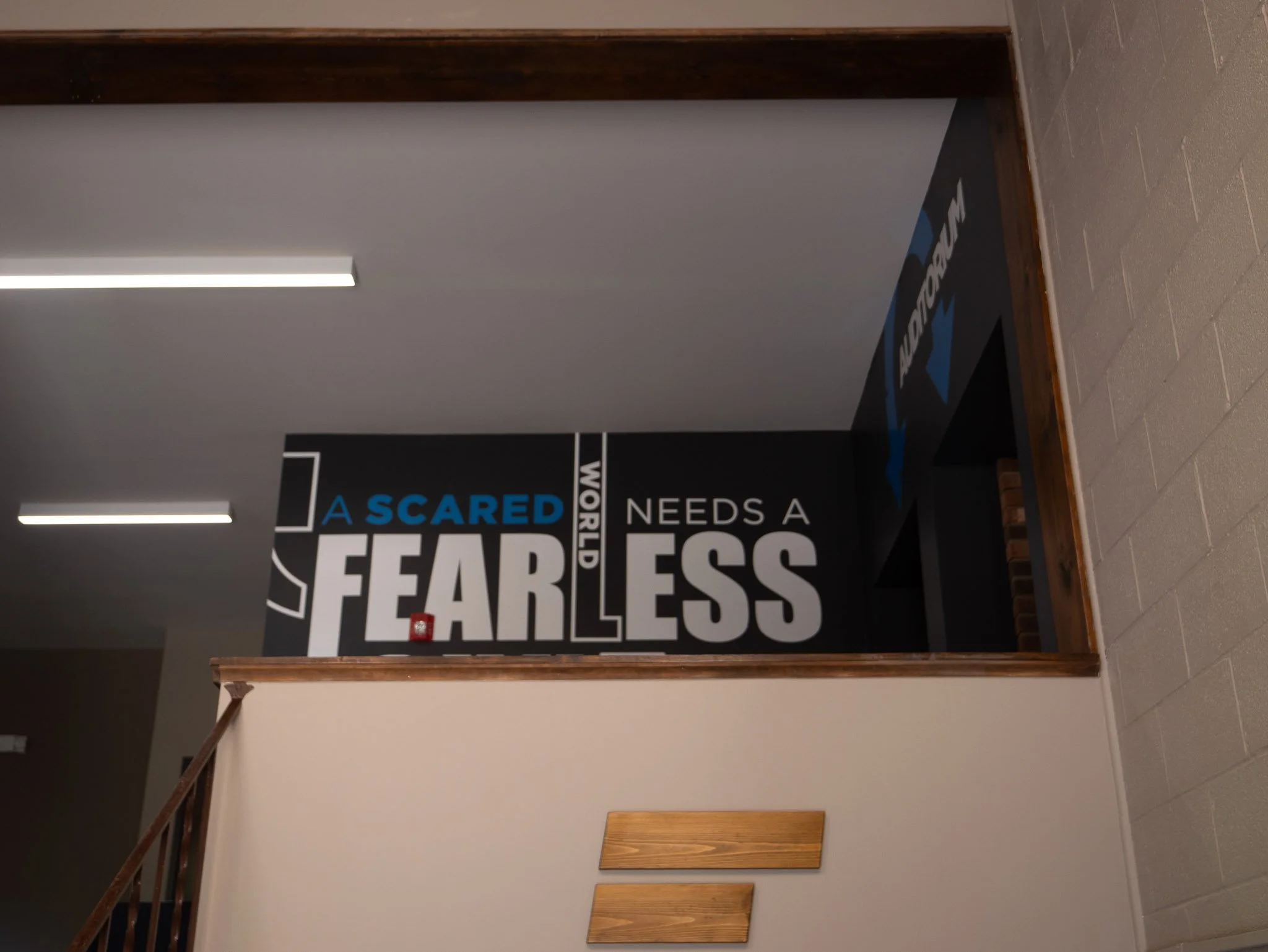 Indoor scene showing a staircase on the left, a wall with a large sign on the right, and ceiling lights. The sign partially reads 'A SCARED' and 'FEARLESS' in bold letters.
