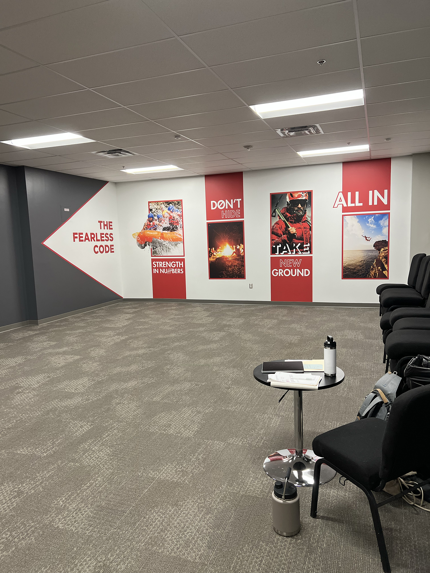 Office waiting area with chairs, a small round table with a water bottle, papers, a laptop, and a backpack. The wall features motivational posters with images of outdoor activities and adventure.