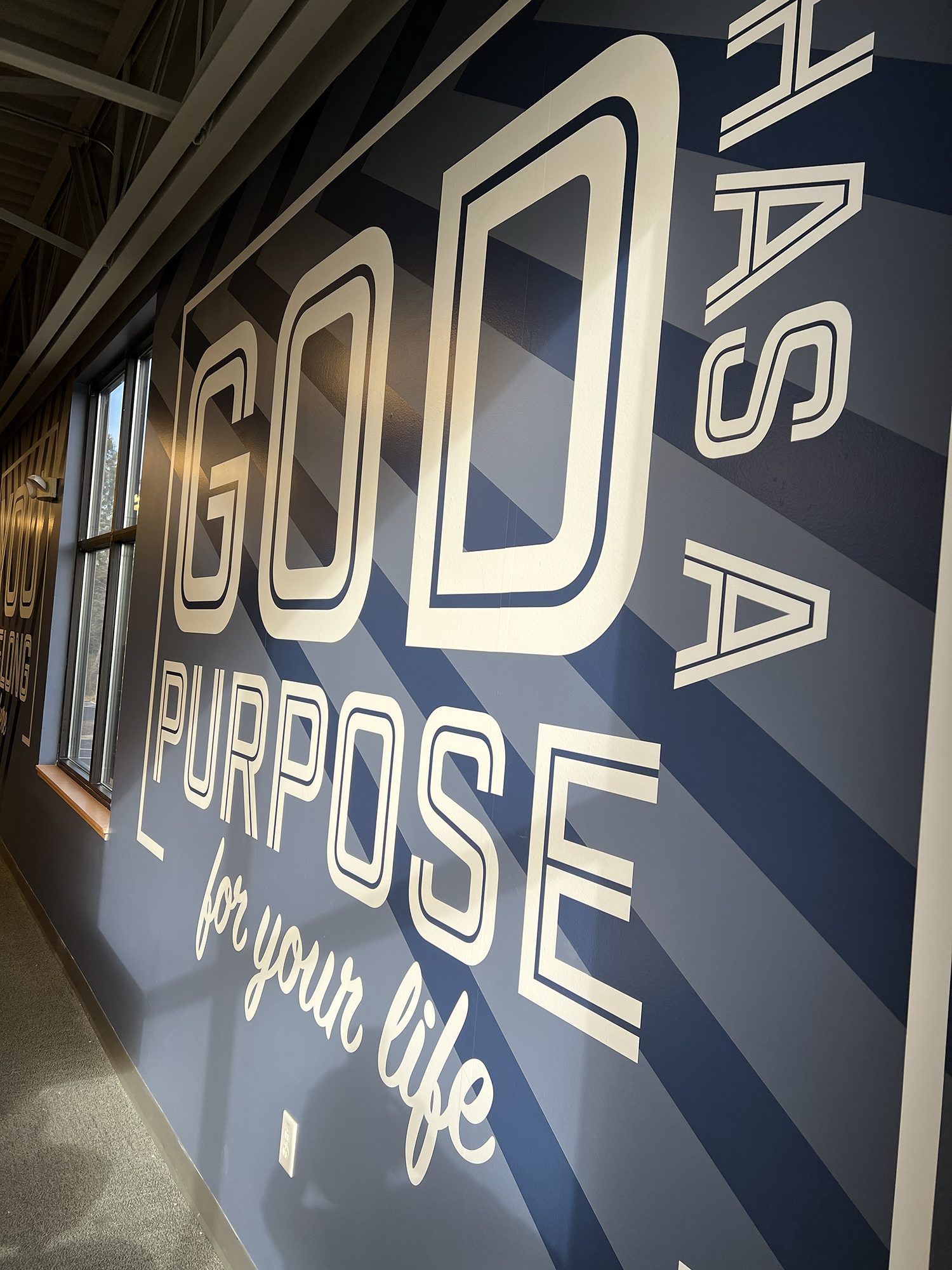 A large interior wall with a blue background and white text reading "LOVE PURPOSE for your life" in a modern, bold font, with a window to the left showing part of the outside scenery.