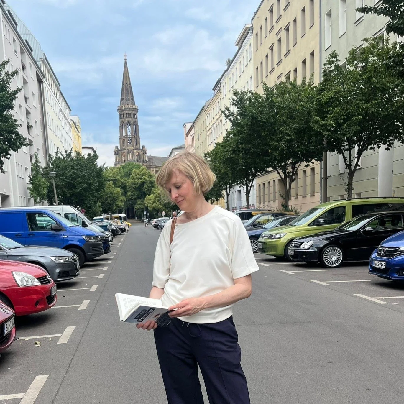 Eine Frau liest ein Buch auf einer Straße mit parkenden Autos, im Hintergrund eine Kirche mit hohem Turm, Bäume und mehrstöckige Gebäude.