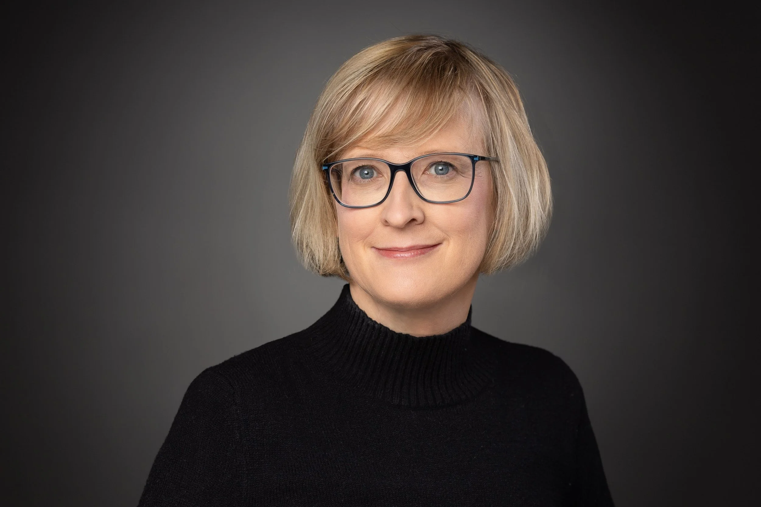 Nahaufnahme Katrin Reiser, mit blonden, kurz gewellten Haaren, schwarzem Rollkragenpullover und Brille vor dunklem Hintergrund.