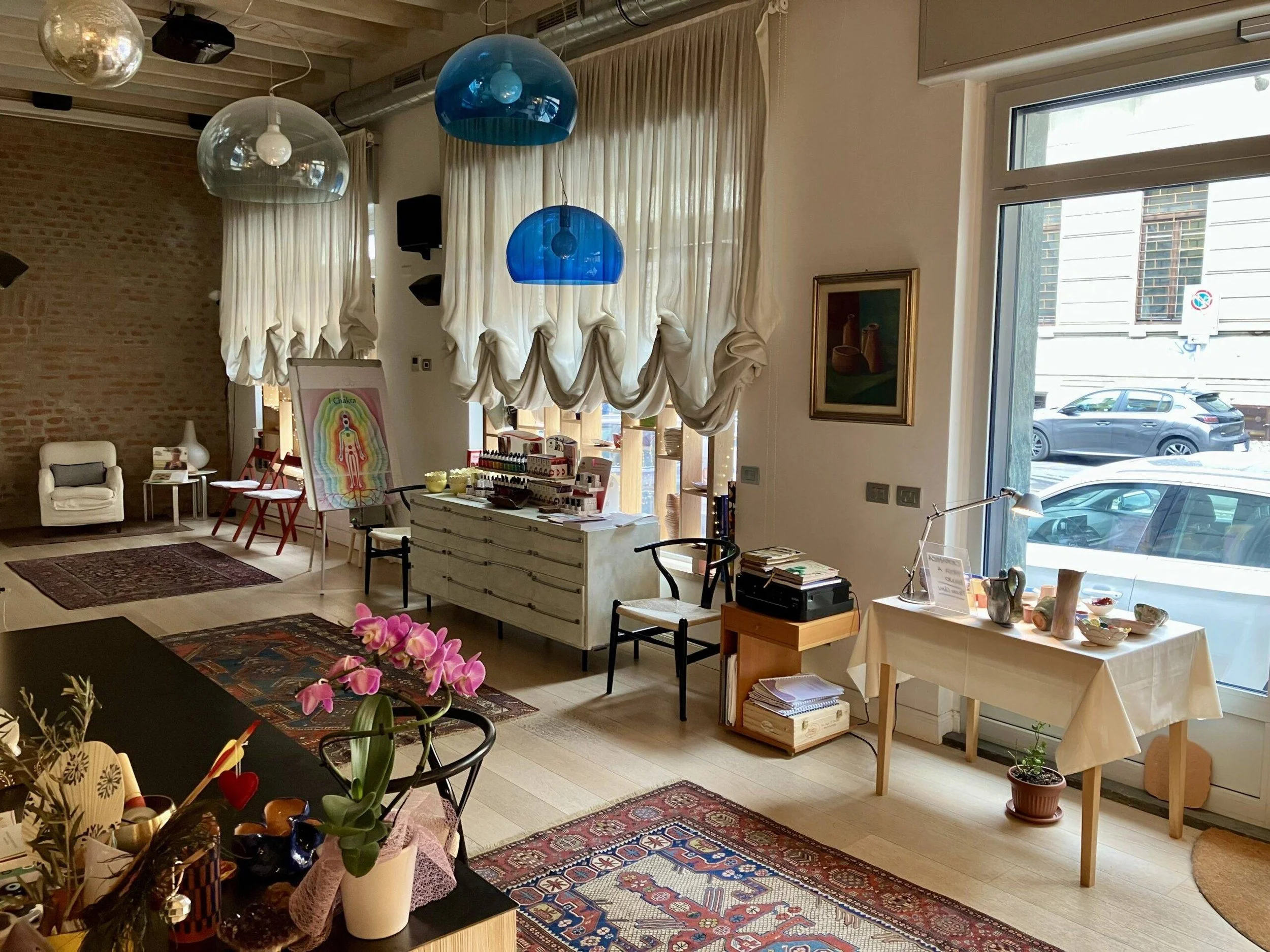 Interno di un negozio con decorazioni e illuminazione sospesa, sedie nere e un tavolo con oggetti, tappeti sul pavimento, grandi finestre con tende chiare, pareti con quadri e esposizione di piante e oggetti decorativi.