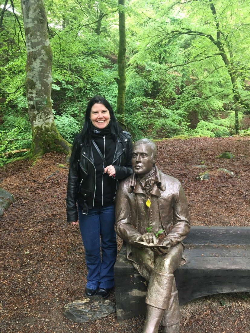 A woman with dark hair wearing a black leather jacket and blue jeans standing next to a bronze statue of a seated man, in a lush green forest setting.